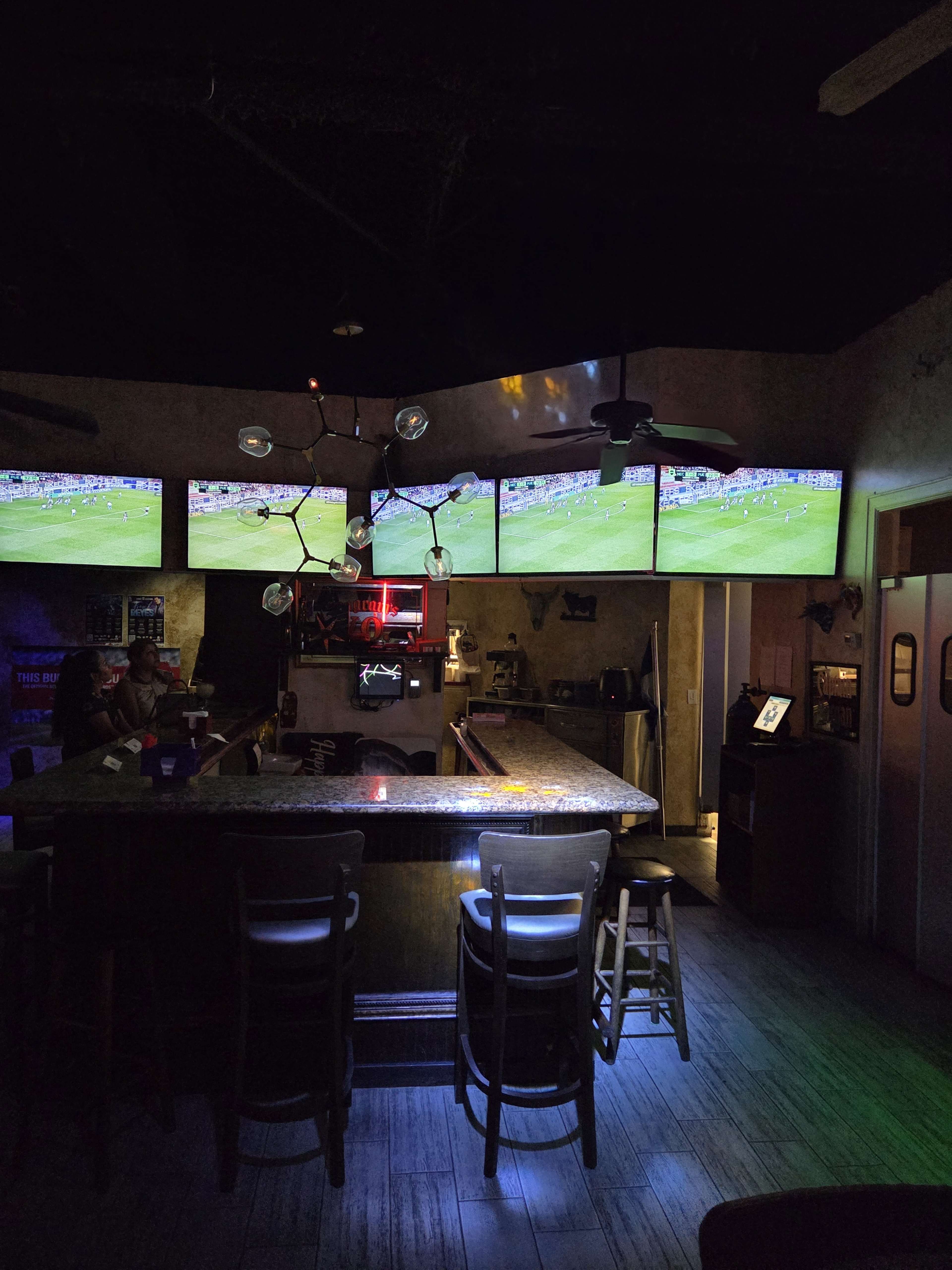 The image shows a dimly lit bar featuring multiple televisions mounted on the wall, displaying a sports game, with a long countertop and a few patrons seated at the bar.