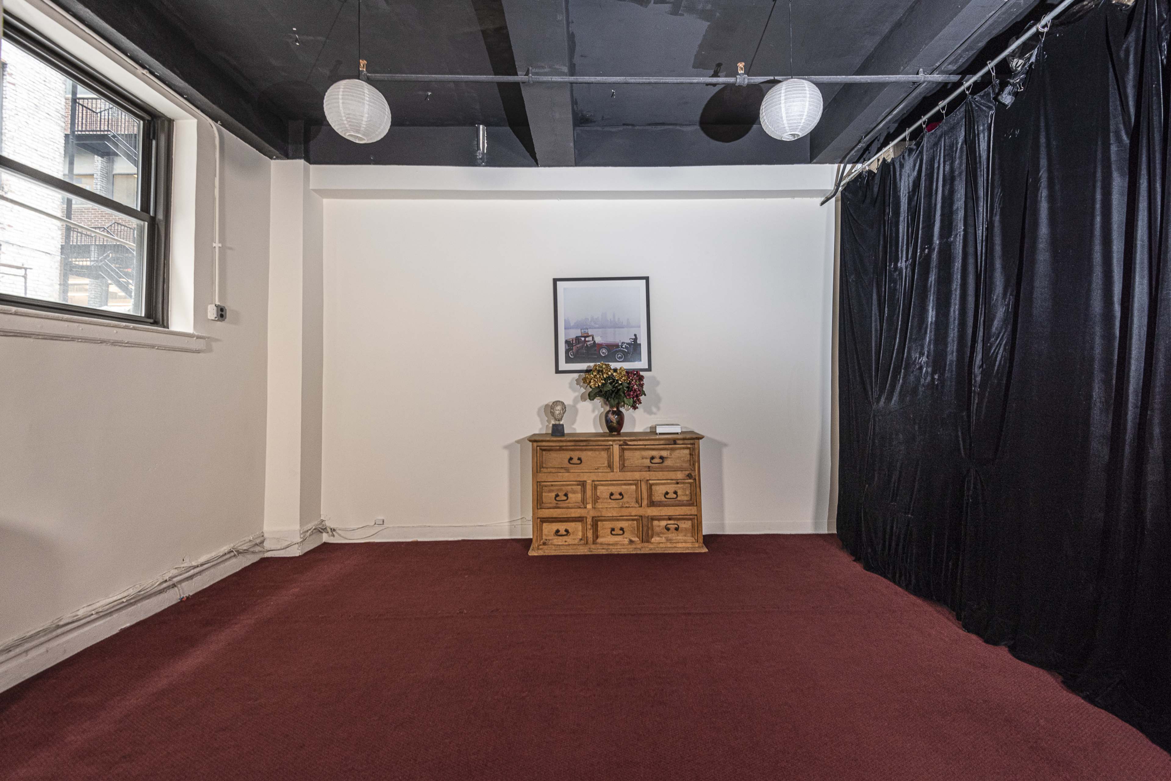 A sparsely furnished room with a red carpet, a wooden dresser, and a wall covered in black drapes, along with a framed picture and a vase of flowers on the dresser.