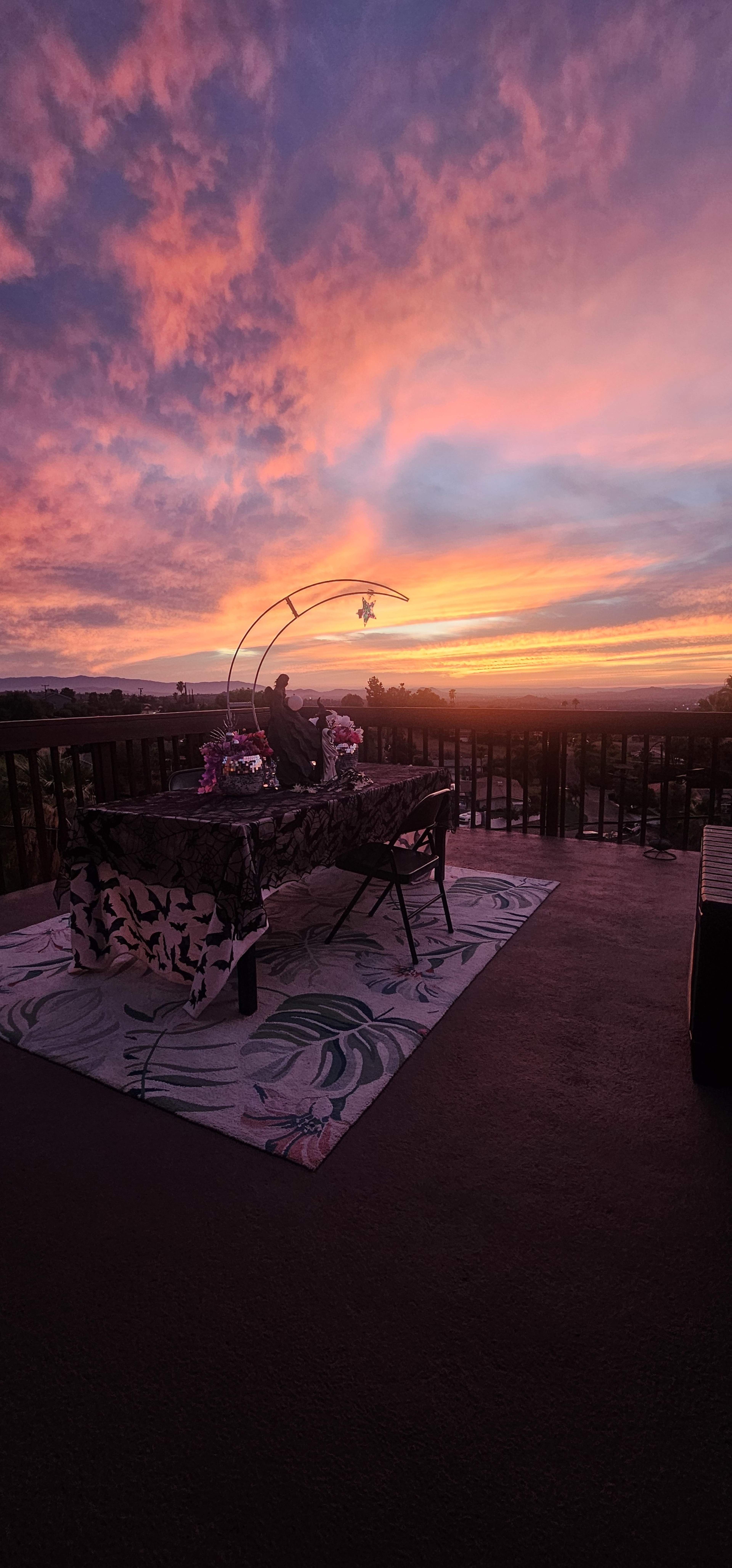 A table adorned with flowers sits on a patterned rug against a backdrop of a vibrant sunset sky.
