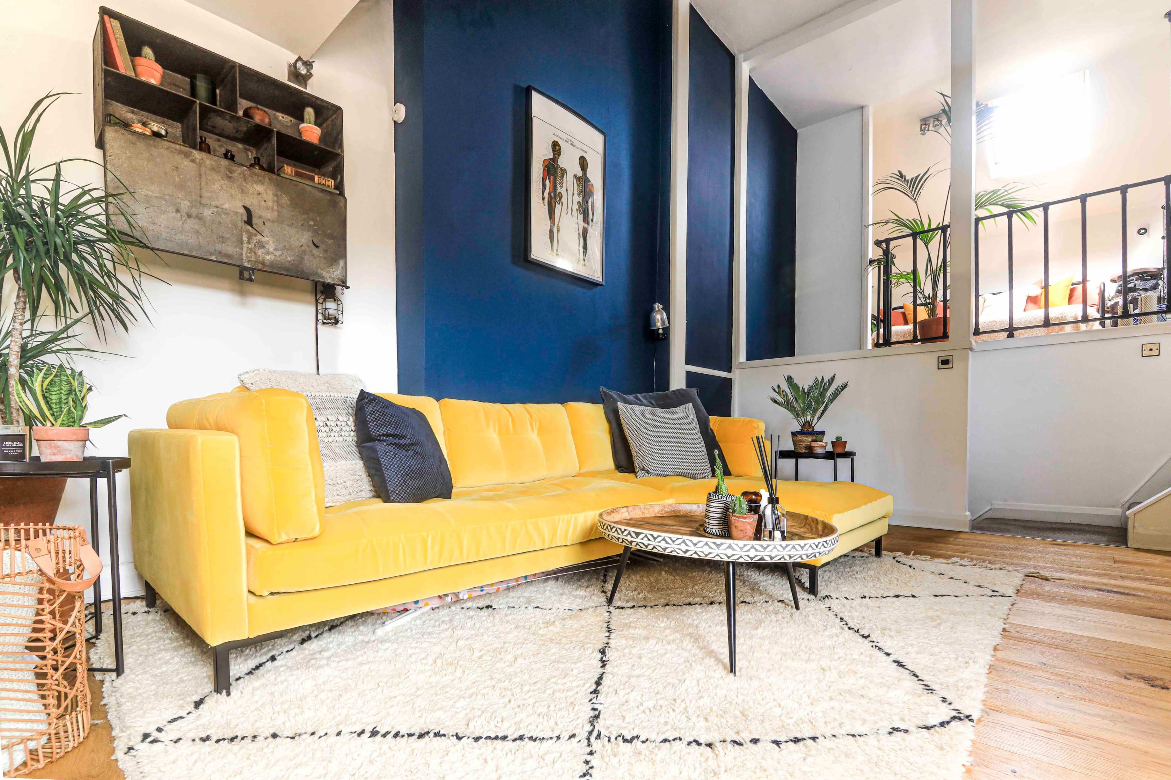 A living room featuring a yellow sofa, a round coffee table, and various plants against a backdrop of blue and white walls.