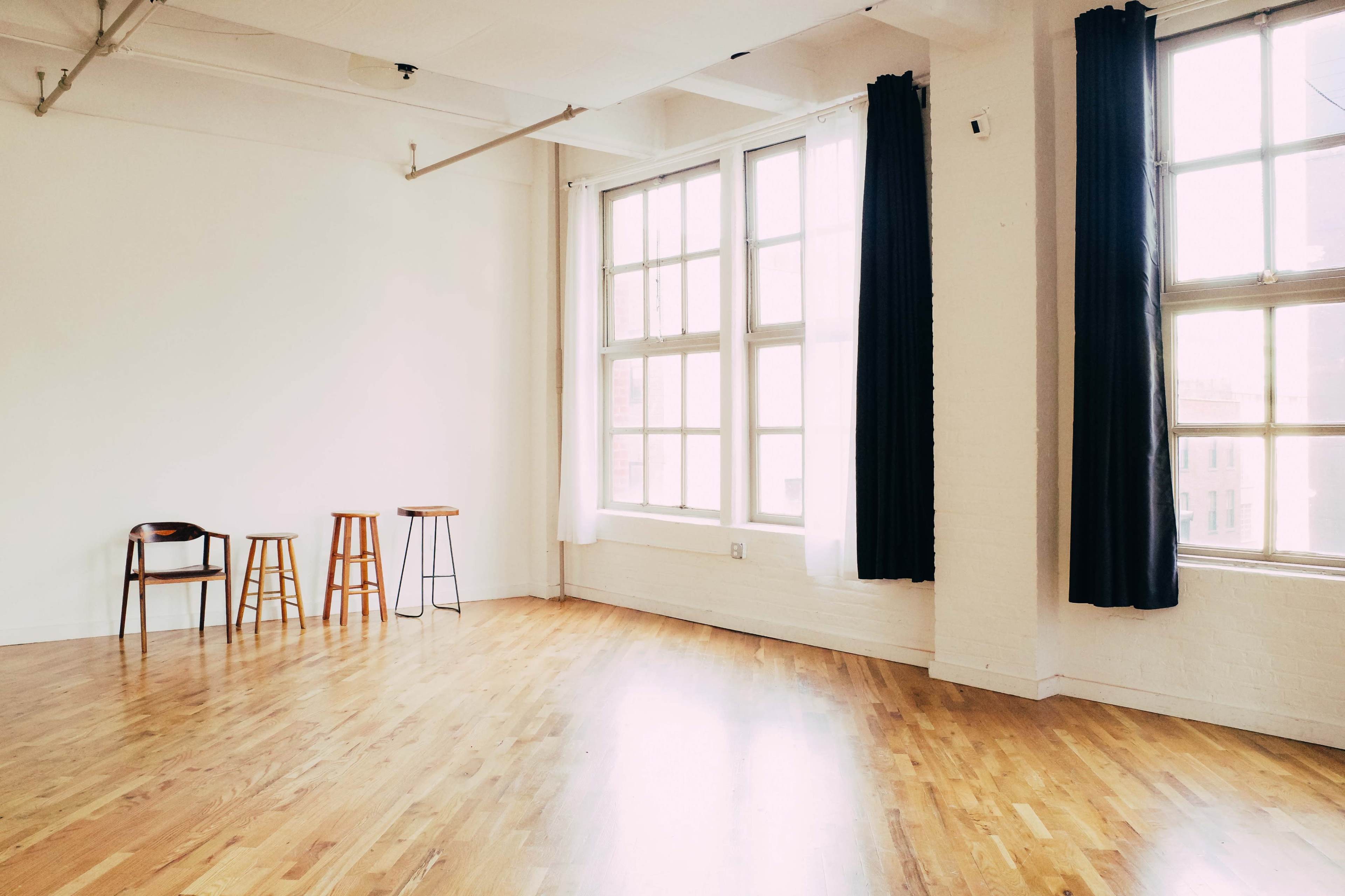 A minimalist room with wooden floors, three stools positioned against a wall, and large windows allowing natural light to illuminate the space.