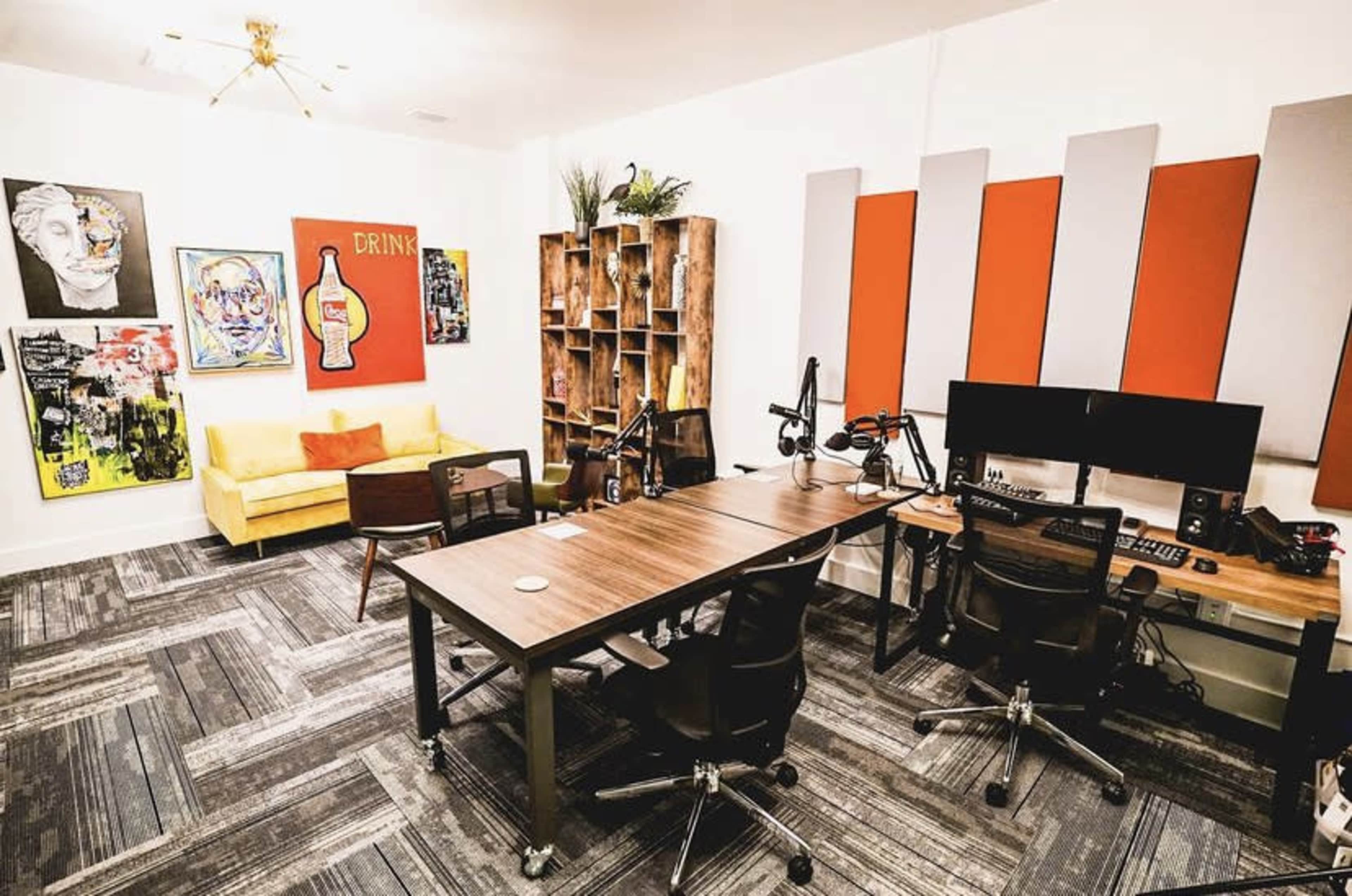 The image shows a modern workspace featuring a sofa, artwork on the walls, a wooden bookshelf, and desks equipped with microphones and computers.