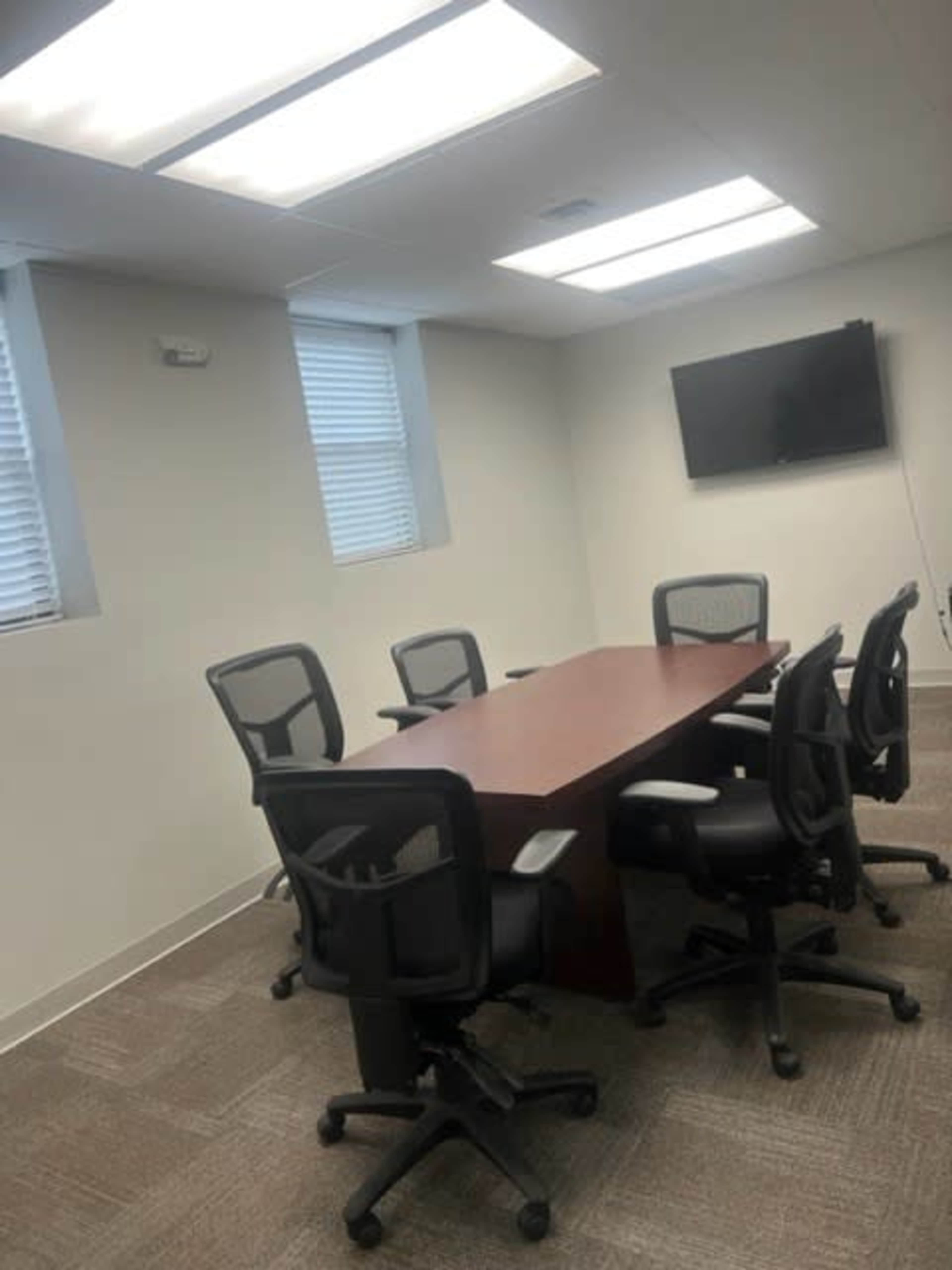 A conference room with a wooden table surrounded by six black office chairs and two windows, featuring a mounted television on the wall.