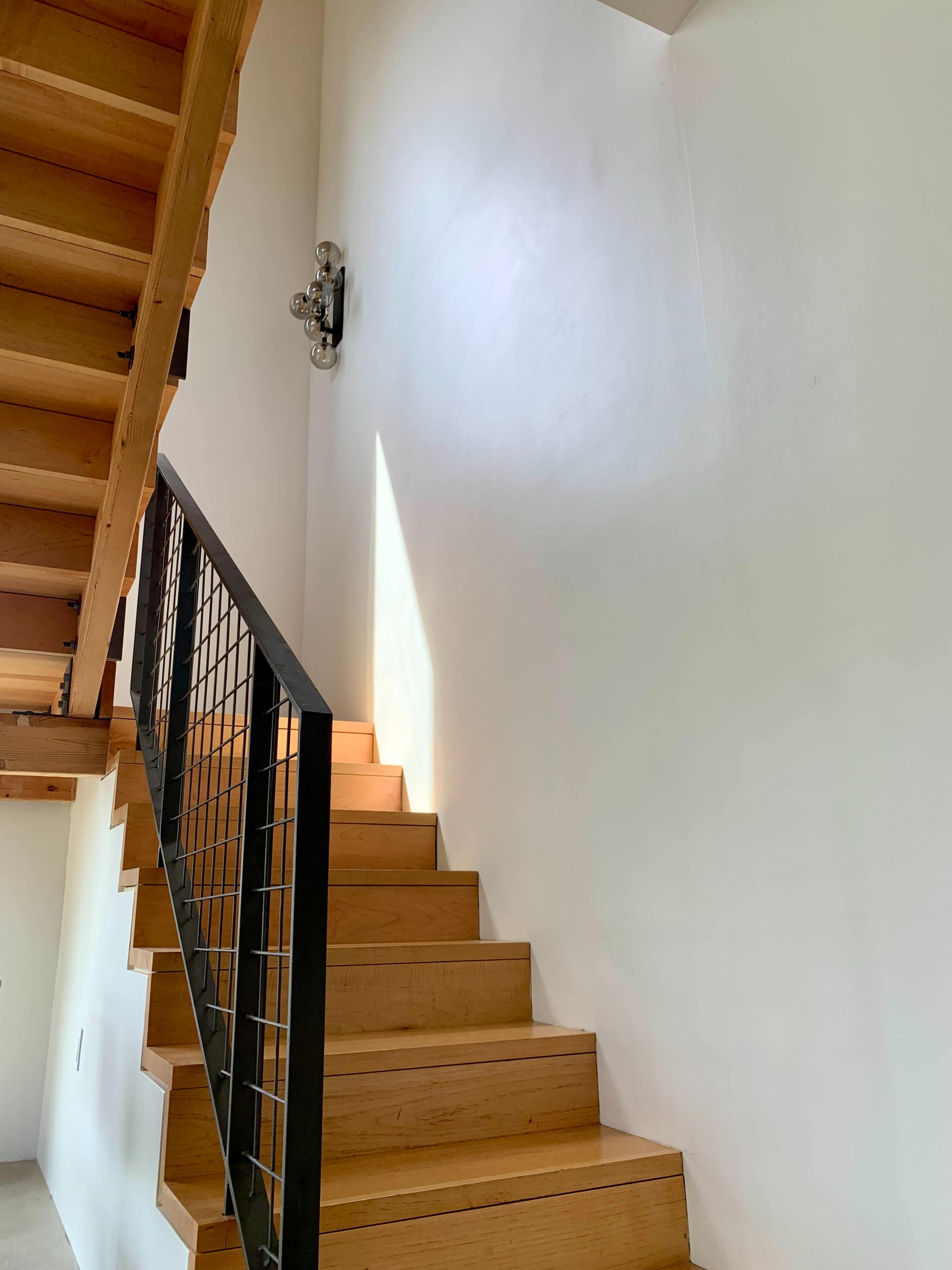 A wooden staircase ascends against a white wall, with a modern light fixture mounted above.