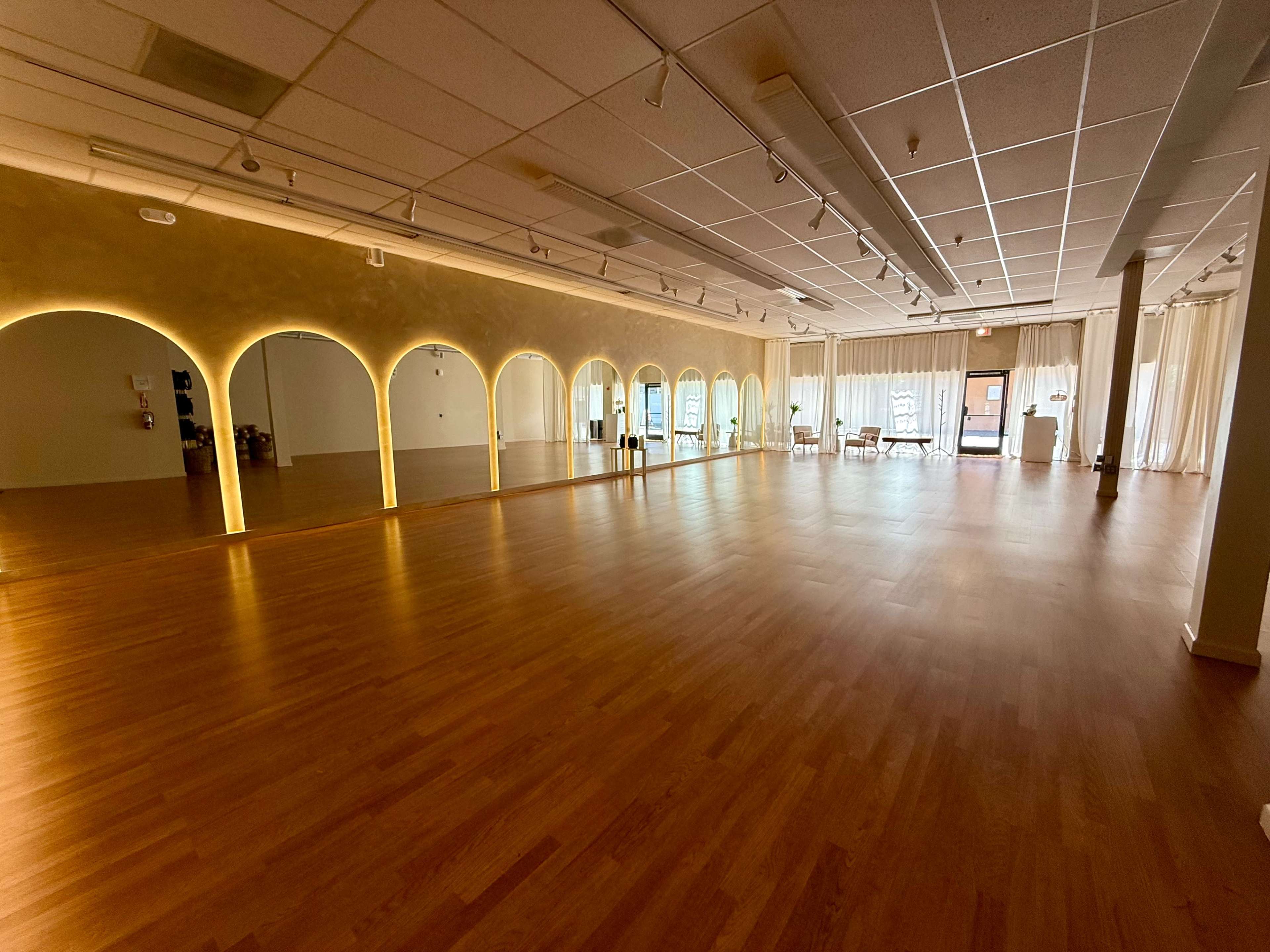 The image shows a spacious, well-lit studio with a wooden floor, large windows, and arches along the walls.