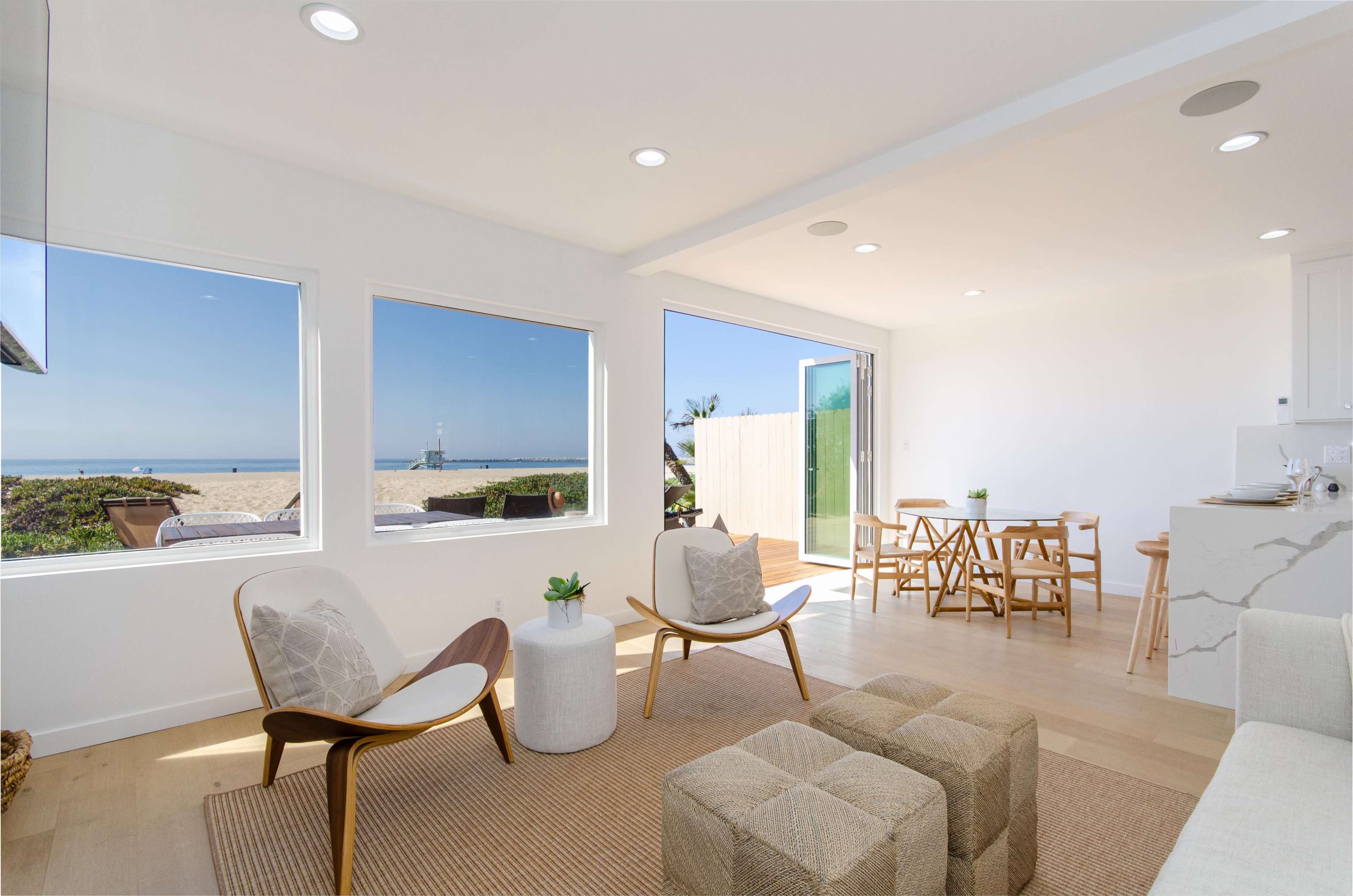 A bright, modern living space with large windows overlooking a sandy beach and ocean, furnished with two chairs, a rug, and a dining area.