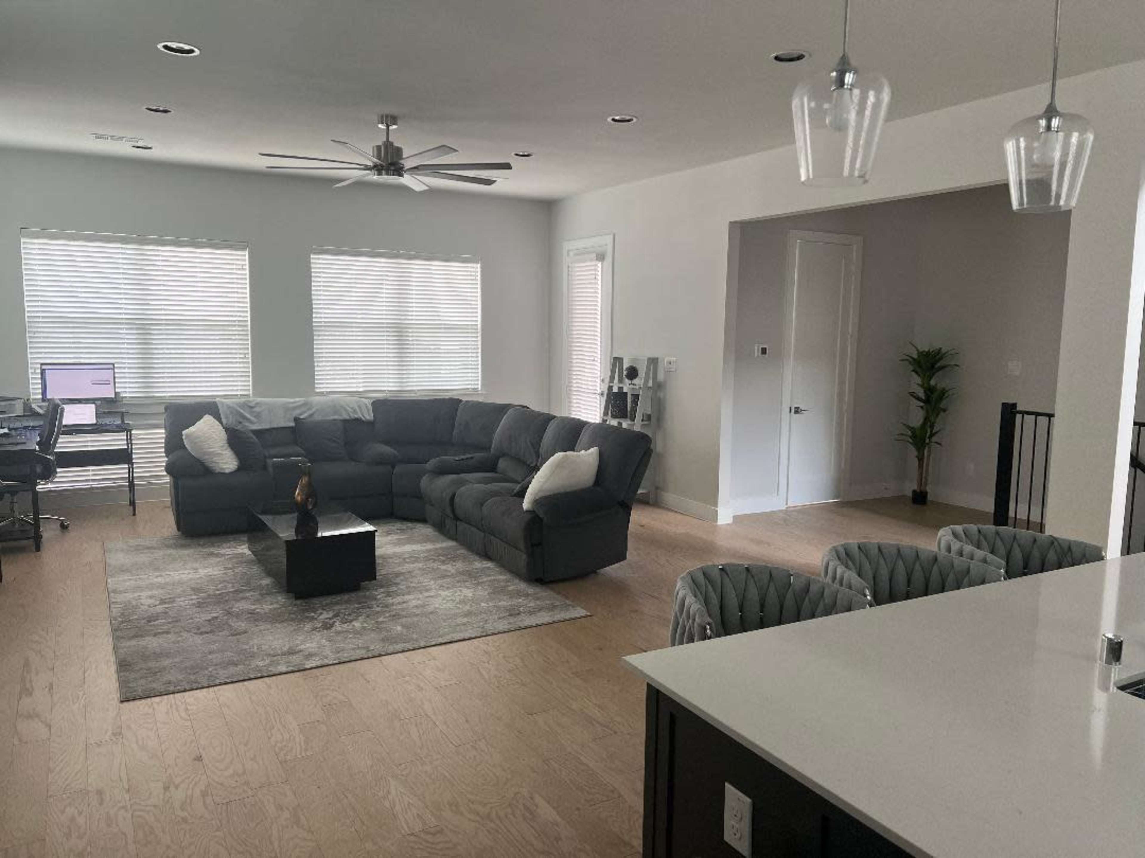 A modern living room features a large sectional sofa, a coffee table, and a desk area with a computer, illuminated by natural light from multiple windows.