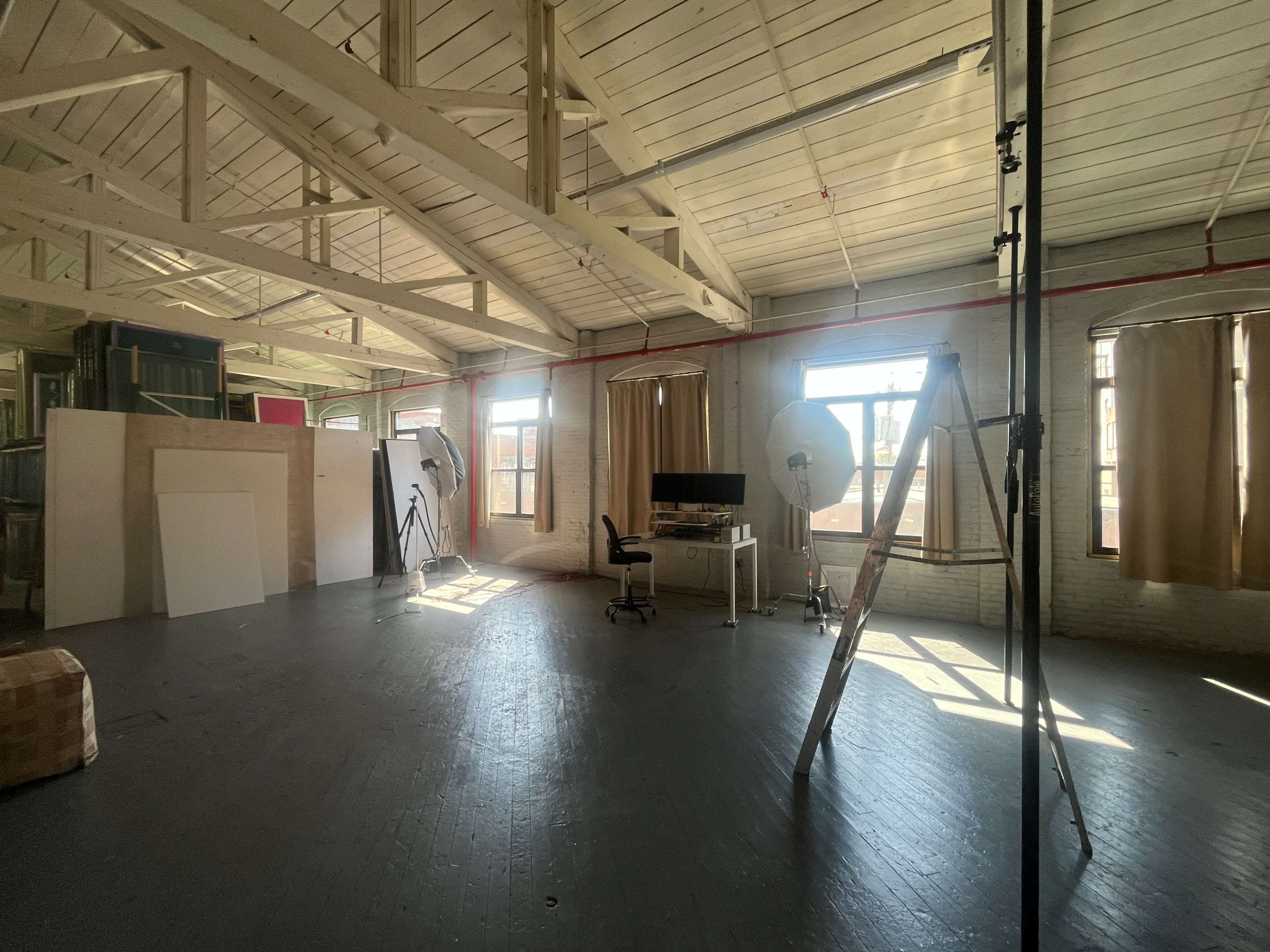 The image shows a spacious studio with high ceilings, large windows, and minimal furnishings, including a desk, chair, and a ladder.