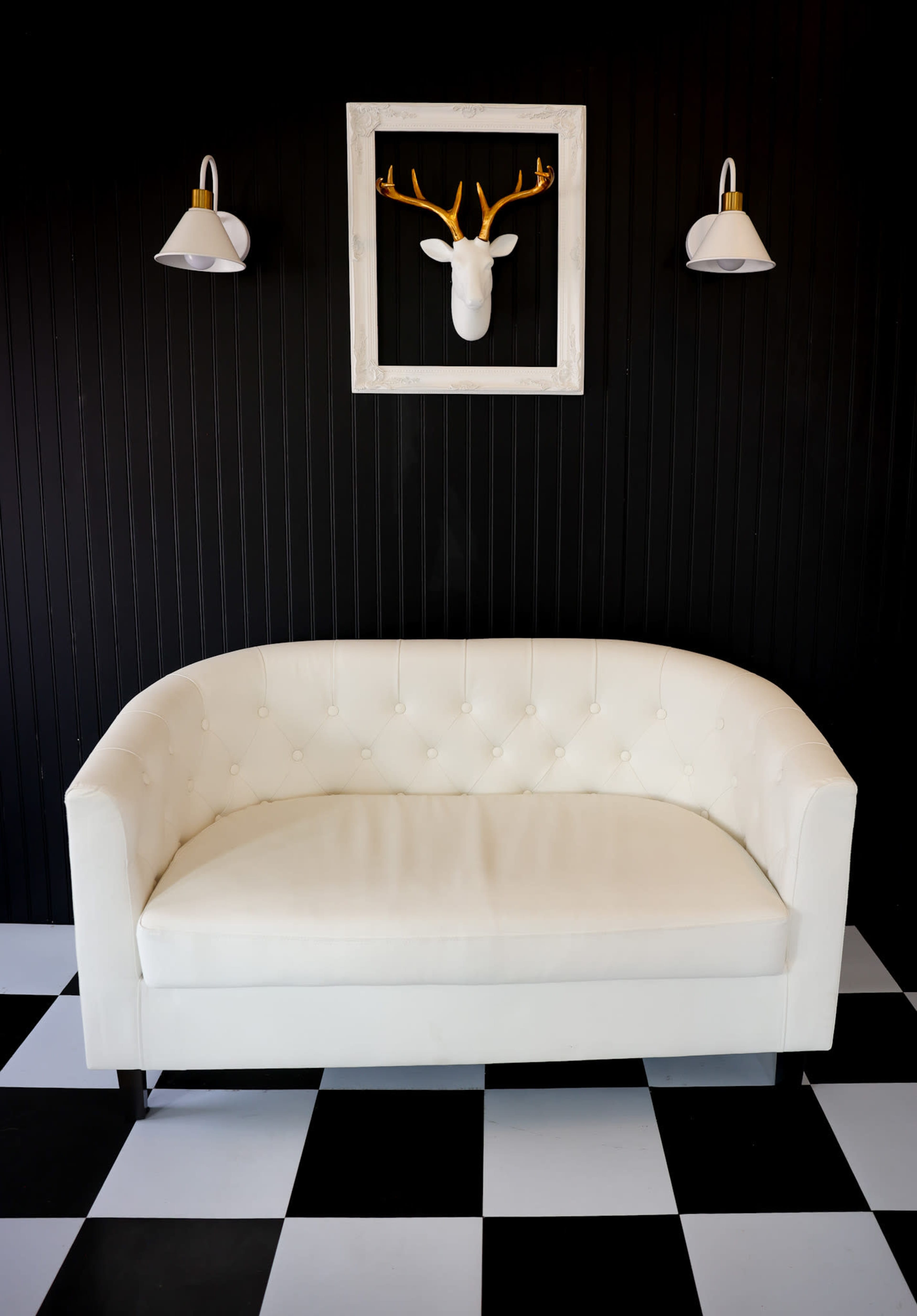 The image features a white tufted sofa in front of a black wall, with a framed deer head mounted above it and two wall sconces on either side.