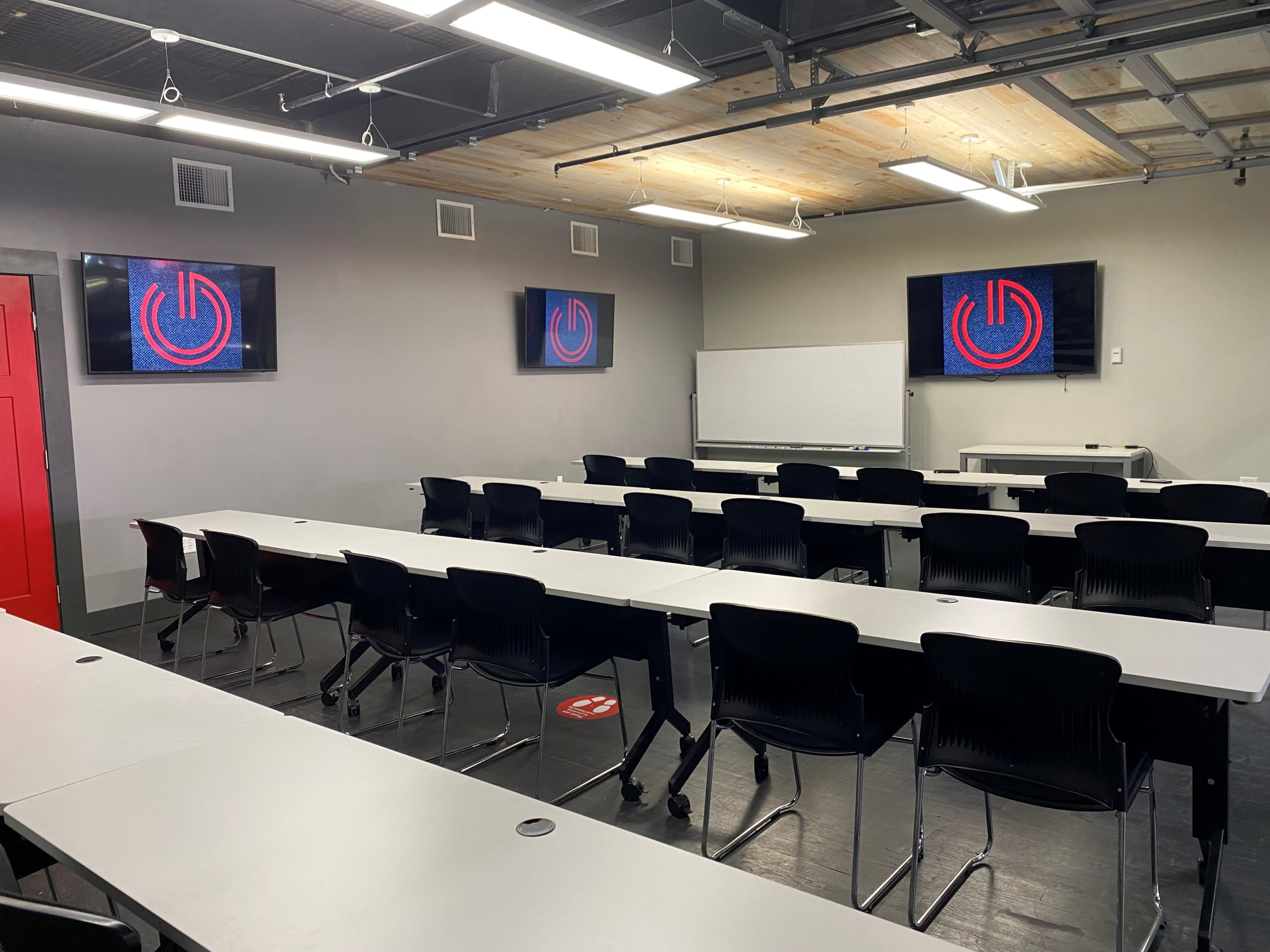 The image shows a modern classroom with multiple rows of tables and black chairs, featuring two screens displaying a circular logo on a gray wall.