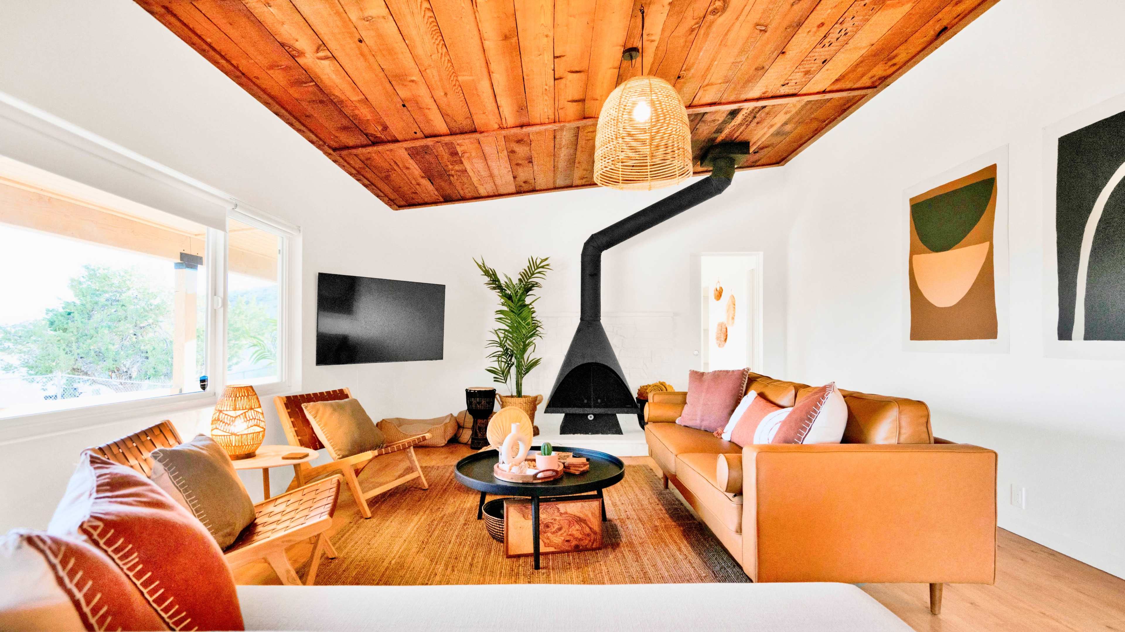 A modern living room features a leather sofa, mid-century chairs, a round coffee table, and a prominent black fireplace, all under a wooden ceiling and accompanied by tropical plants.