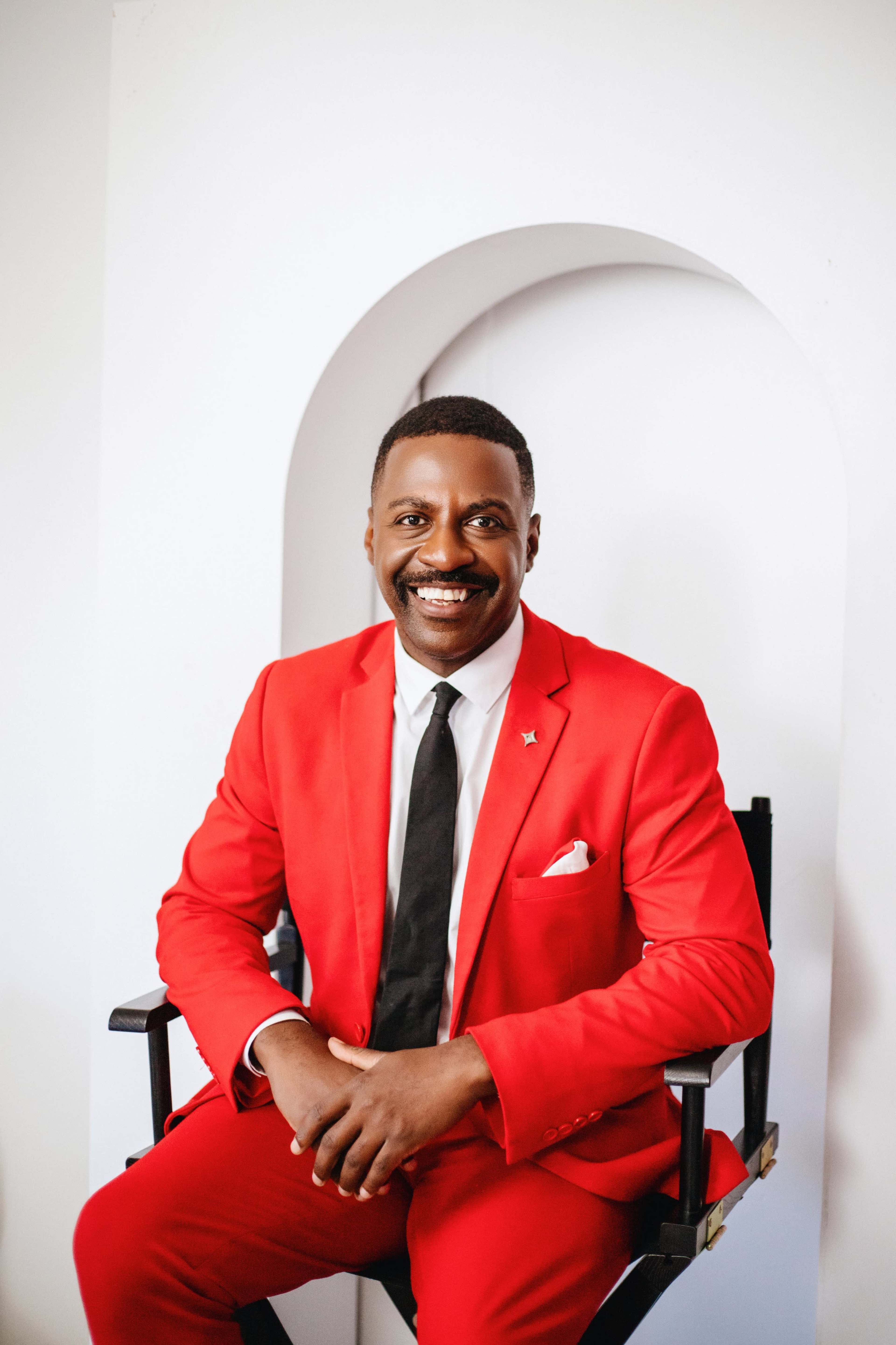 A man in a bright red suit sits on a director's chair, smiling and facing the camera.