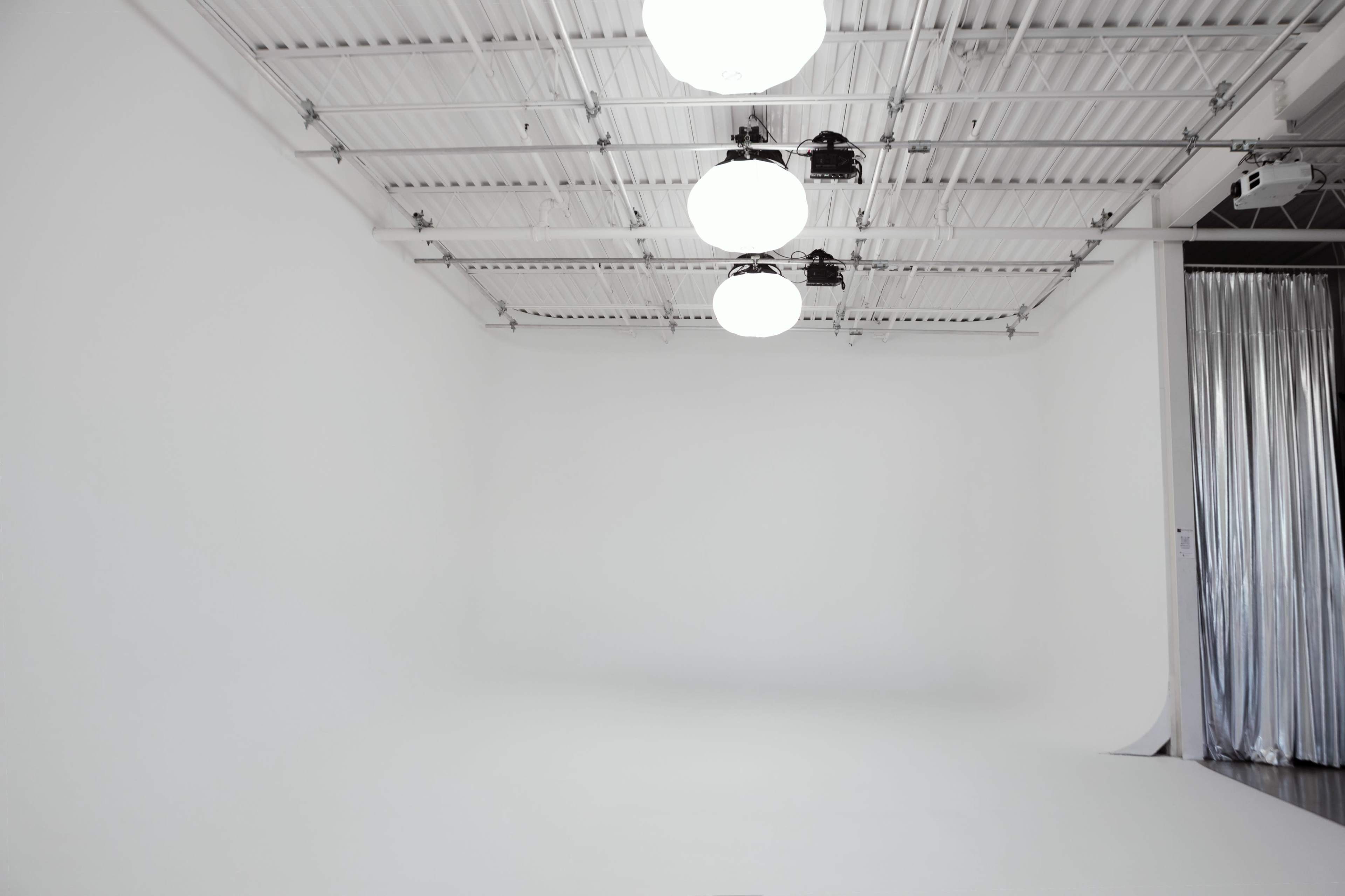 The image shows a spacious, empty studio with white walls and three round light fixtures hanging from the ceiling.