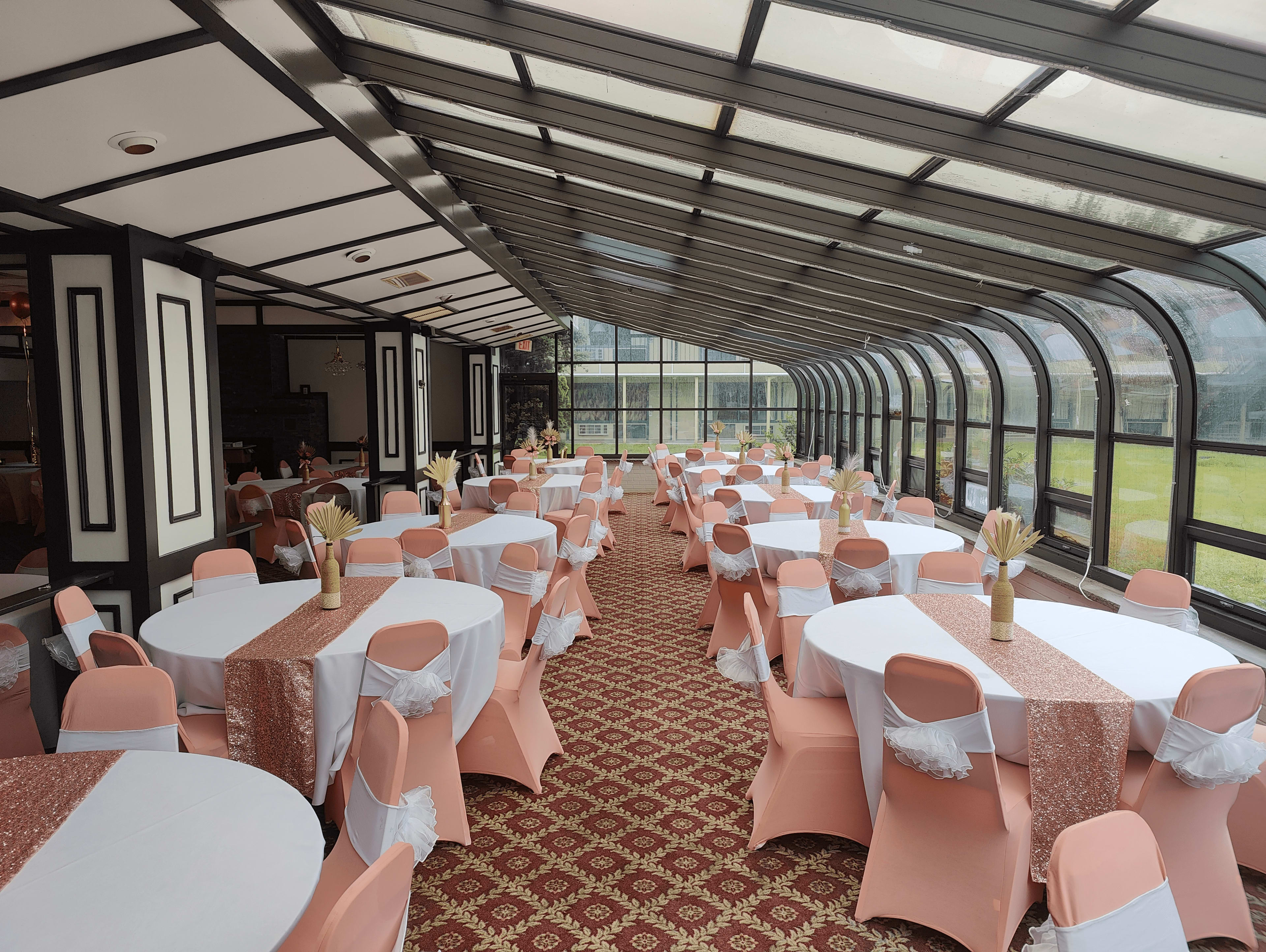 A banquet hall with round tables covered in white cloths and pink chair covers, arranged under a glass roof with large windows overlooking a green area.