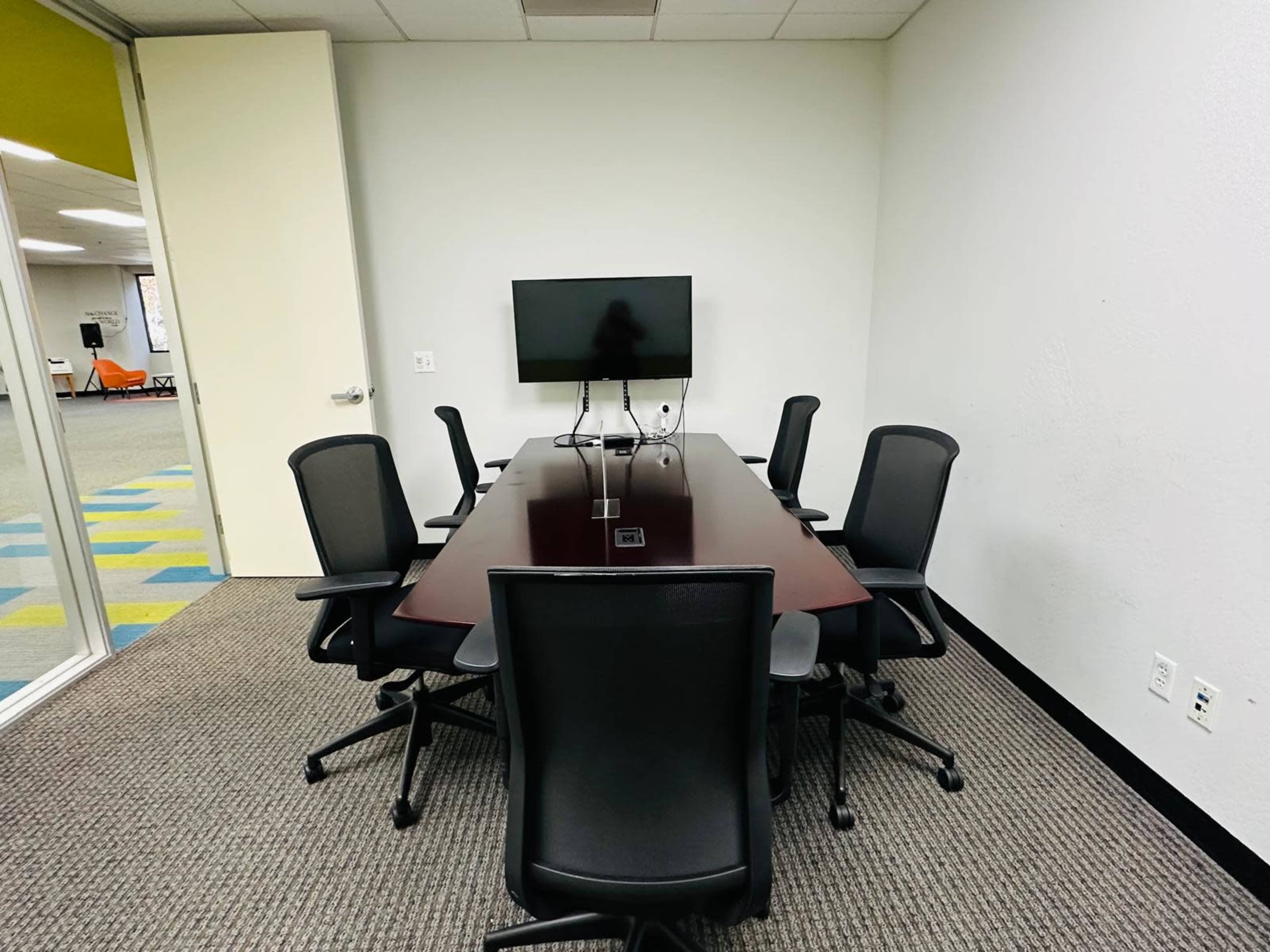 A conference room contains a long table surrounded by six office chairs, with a mounted television on the wall.