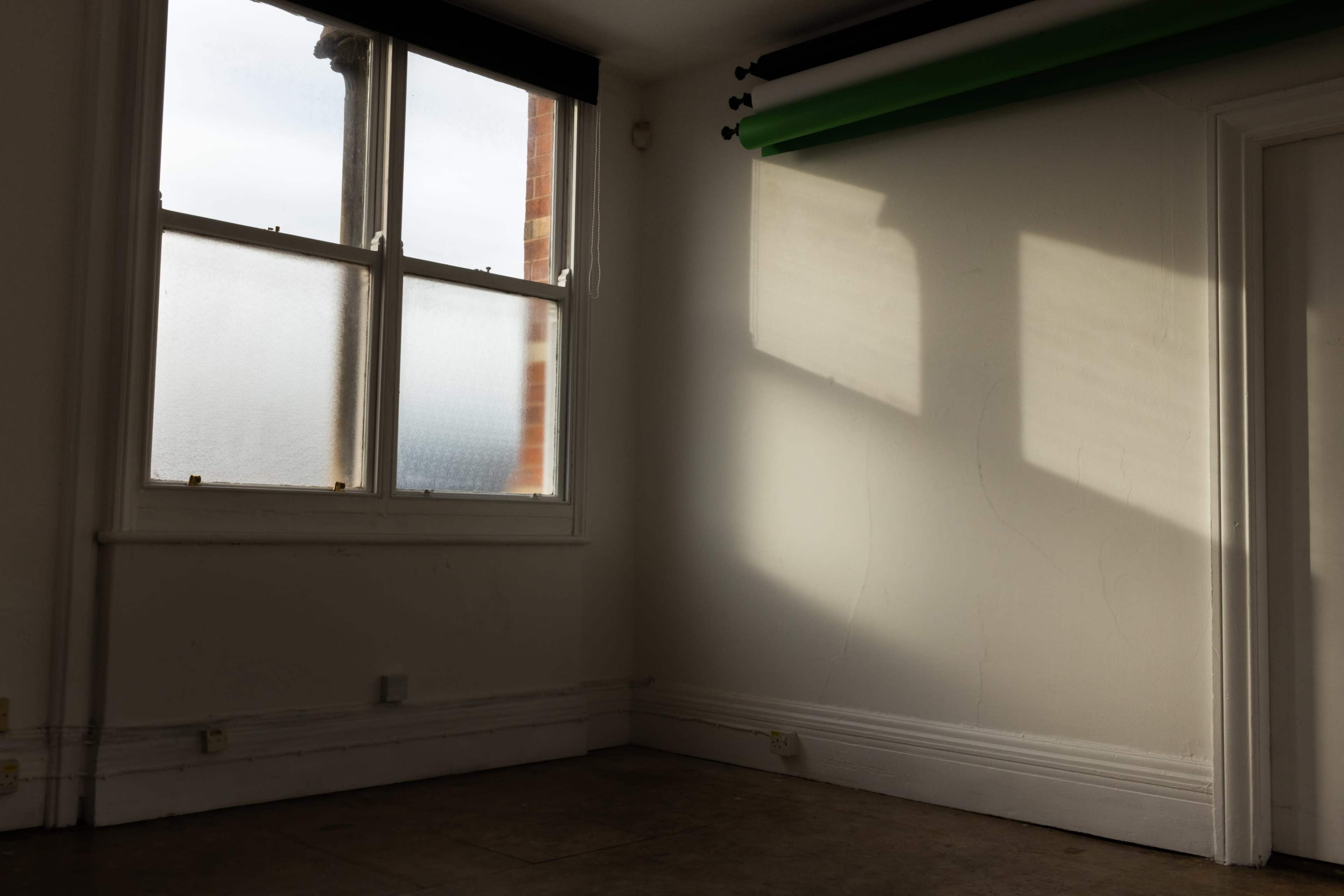 A sunlit room with a large window and a green backdrop roll hanging on the wall.