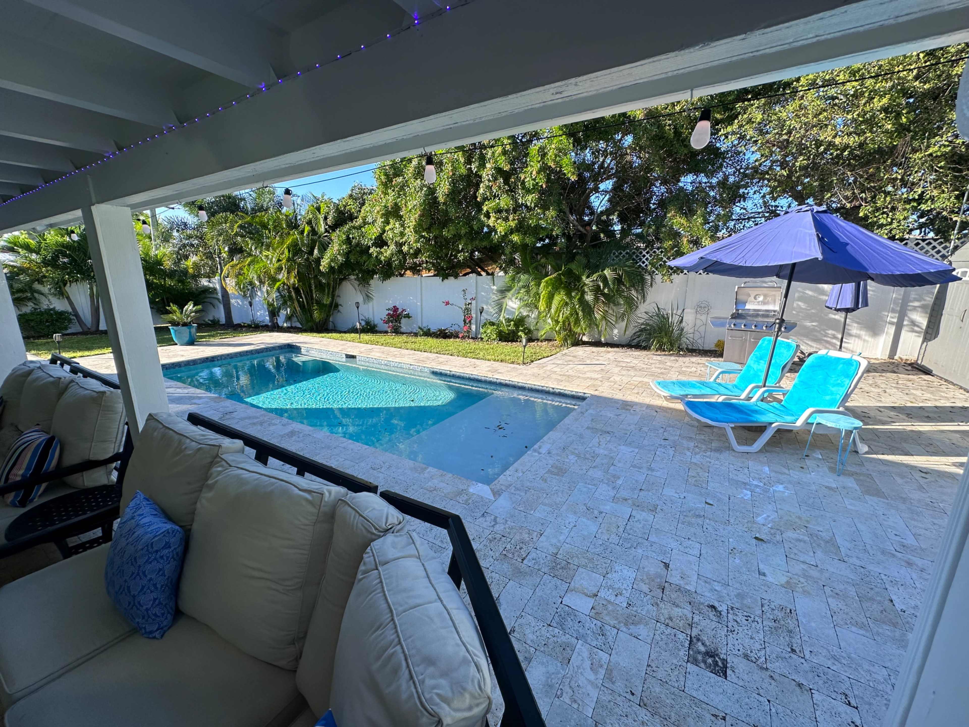 The image shows a backyard with a swimming pool, two blue lounge chairs, a patio area with a couch, and a large umbrella.