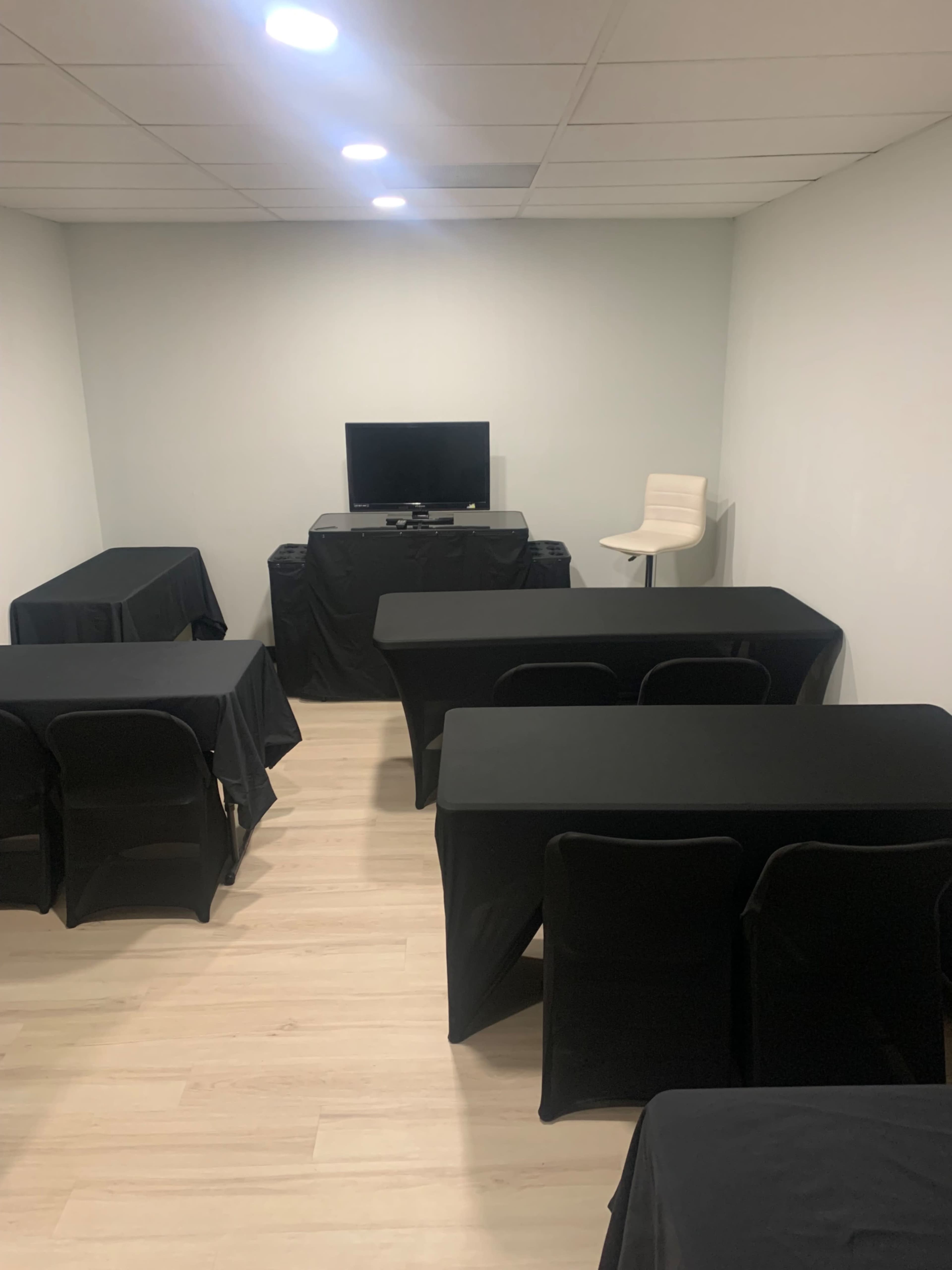 The image shows a small meeting room with several black tables and chairs arranged around a television on a stand.