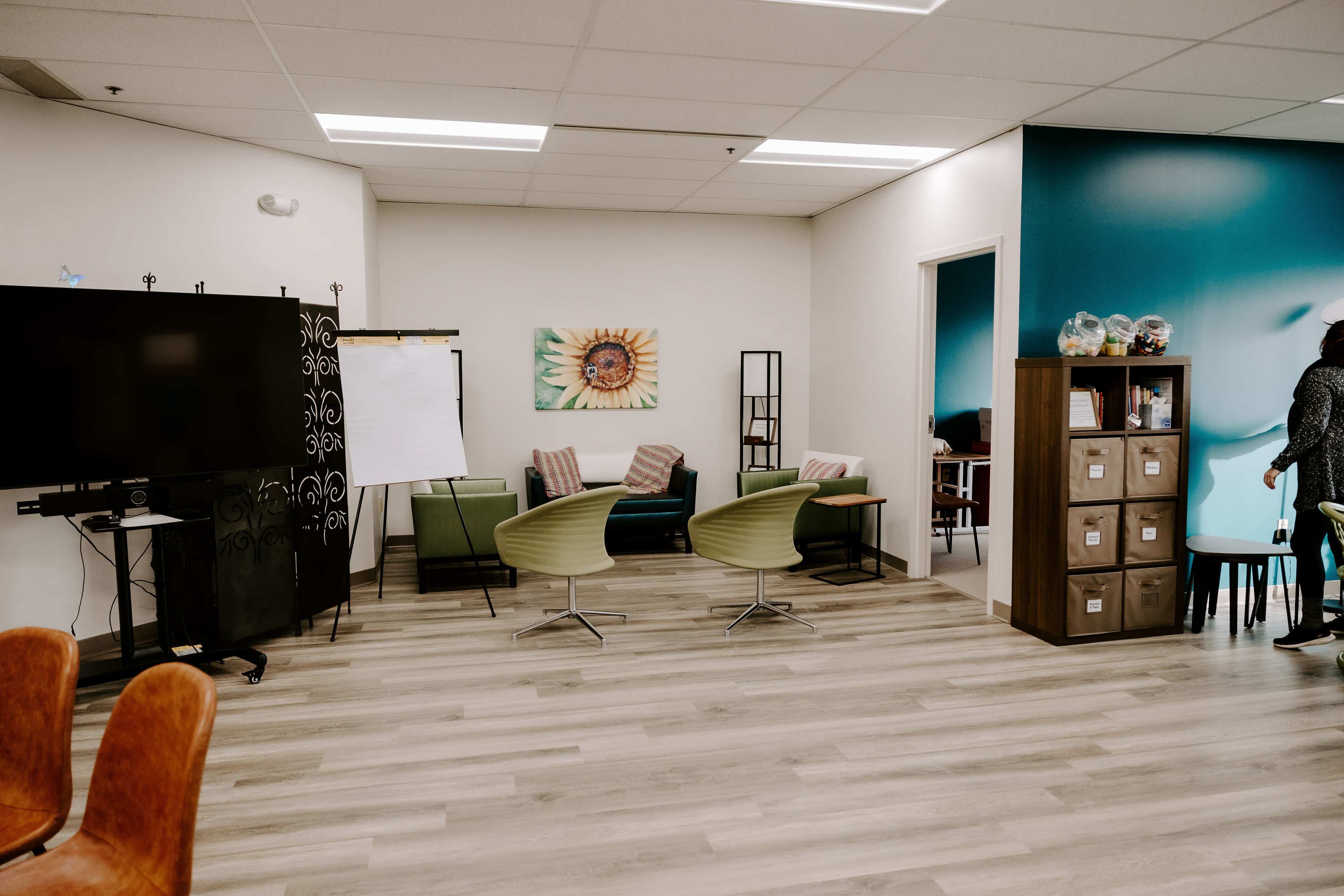 The image depicts a modern office space with seating areas, a large television, and a wall featuring a sunflower painting.