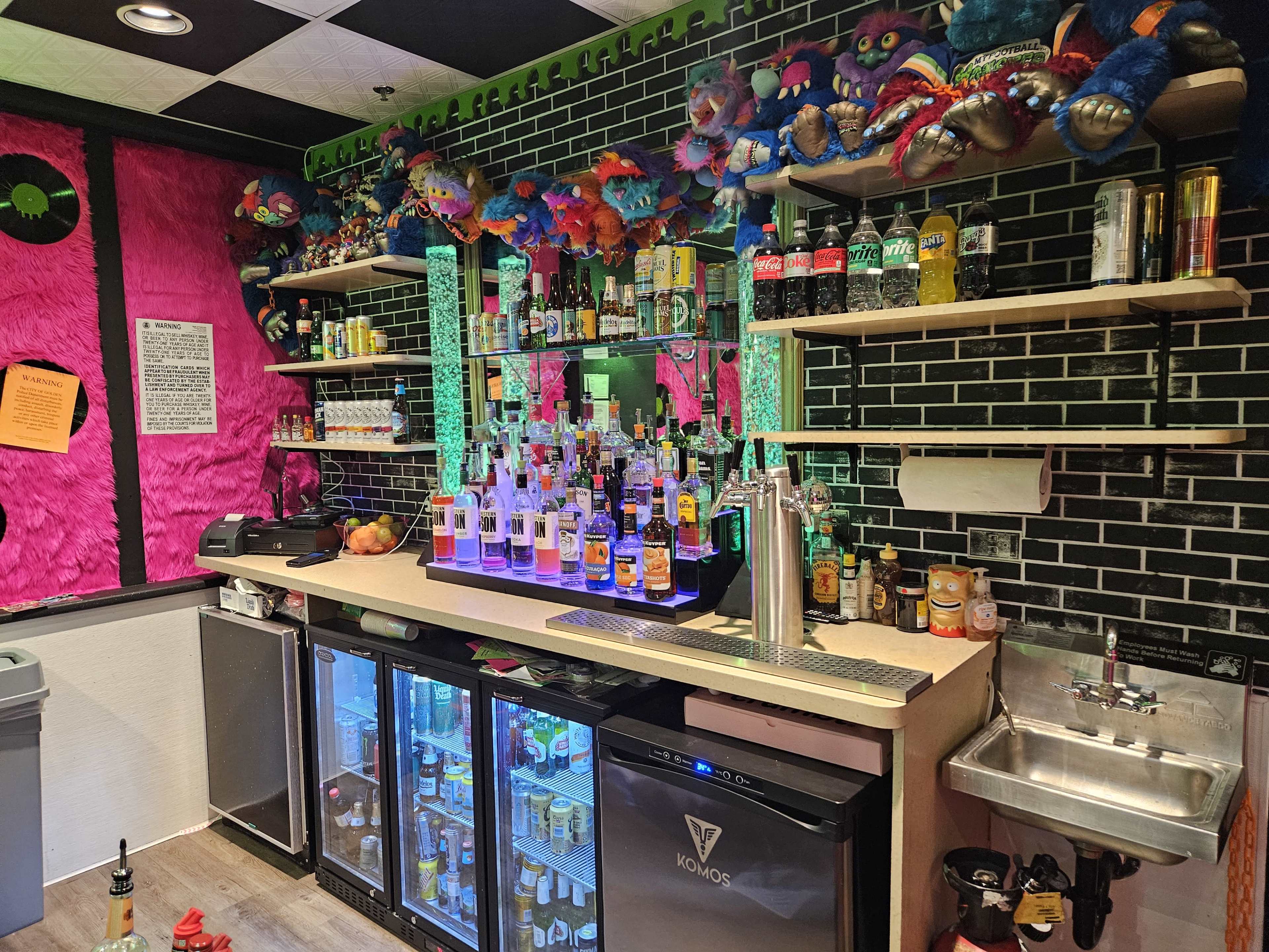 The image shows a colorful bar area with a variety of alcoholic beverages displayed on shelves against a black-tiled wall, featuring neon lighting and decorative elements.