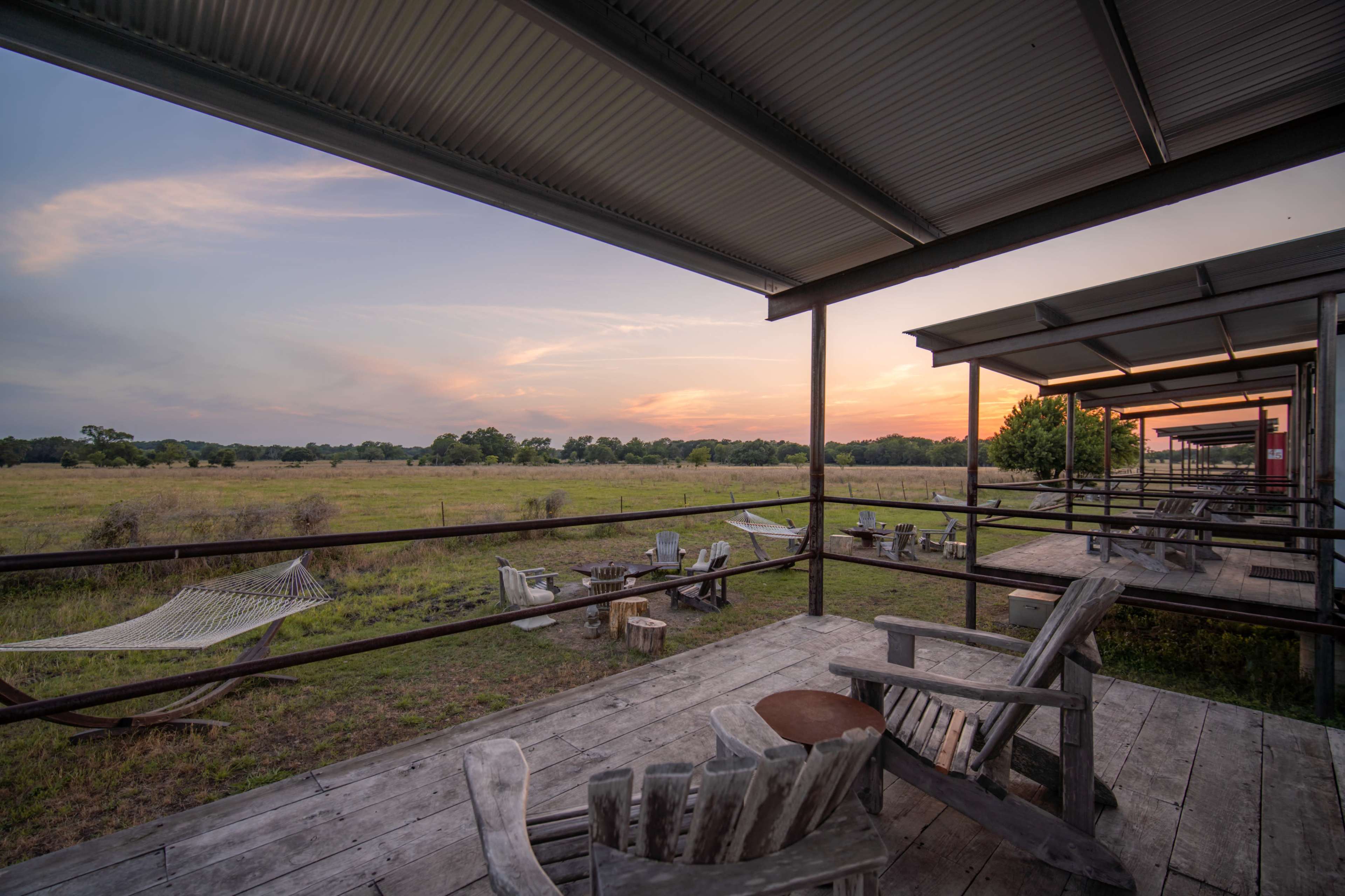 A wooden deck with several chairs overlooks a grassy field under a colorful sunset sky.