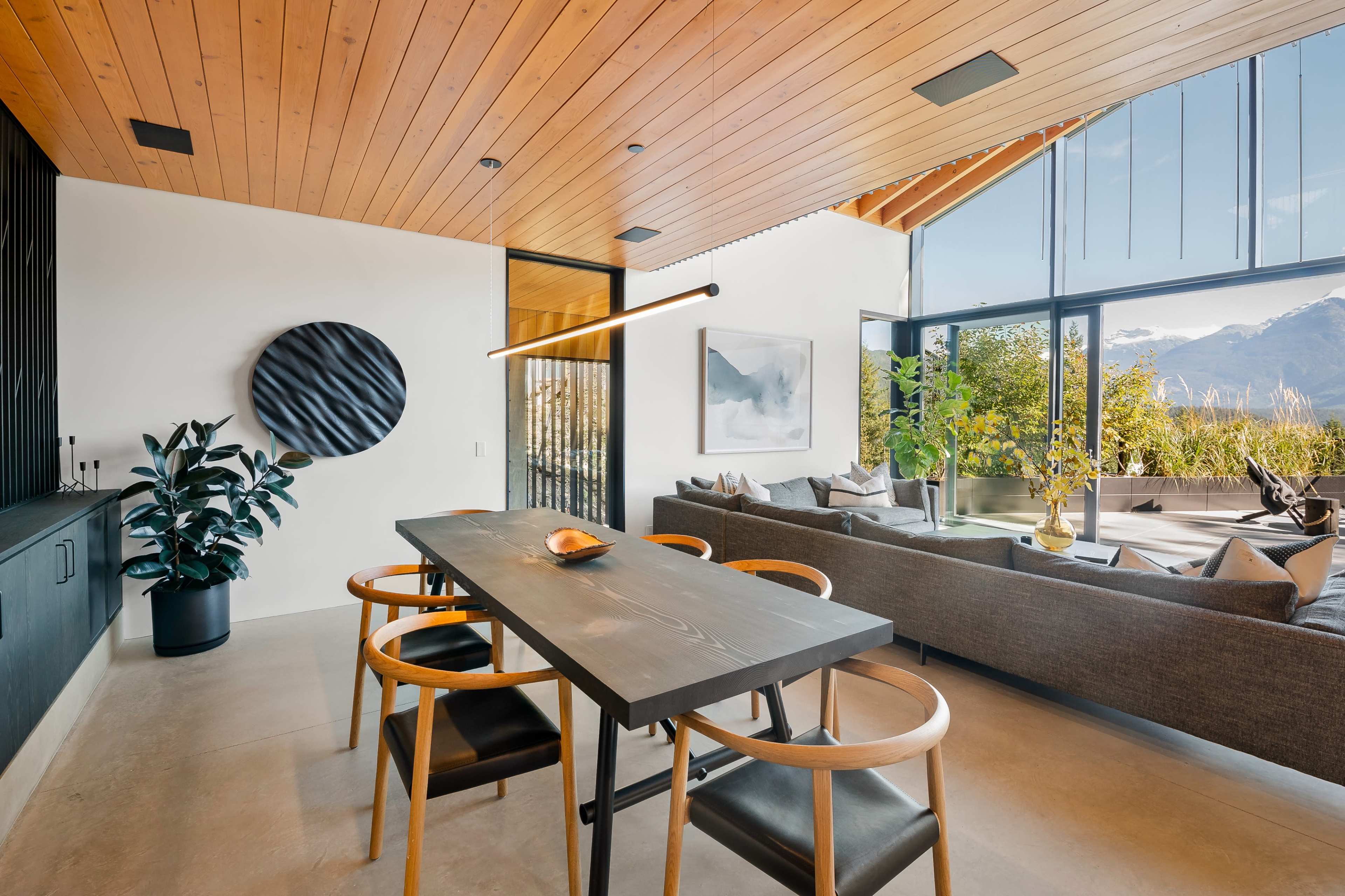 A modern dining area features a black table with wooden chairs, surrounded by large windows that overlook a scenic outdoor space.