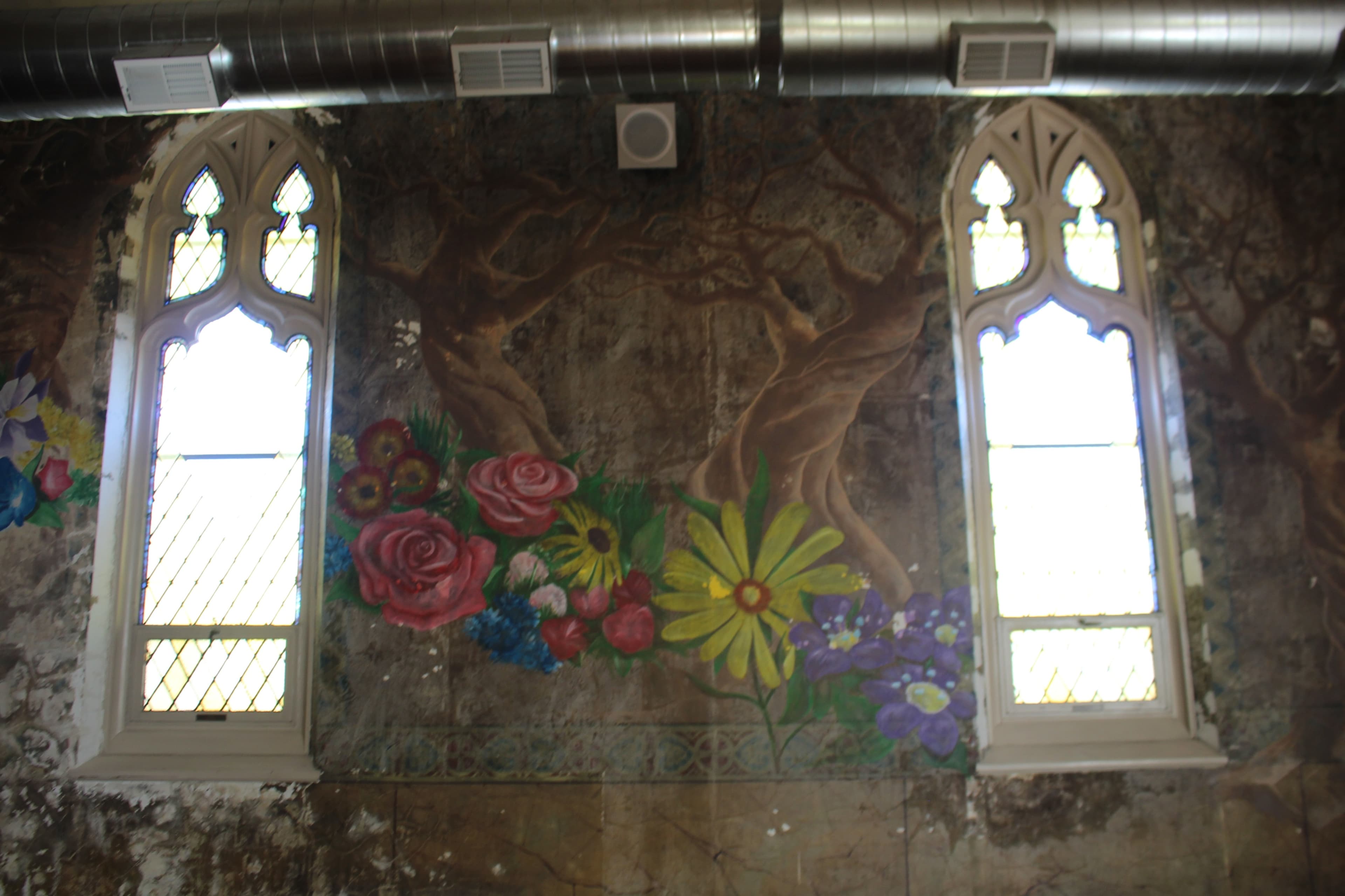 A wall with a faded mural featuring flowers and trees, flanked by arched windows with decorative glass.