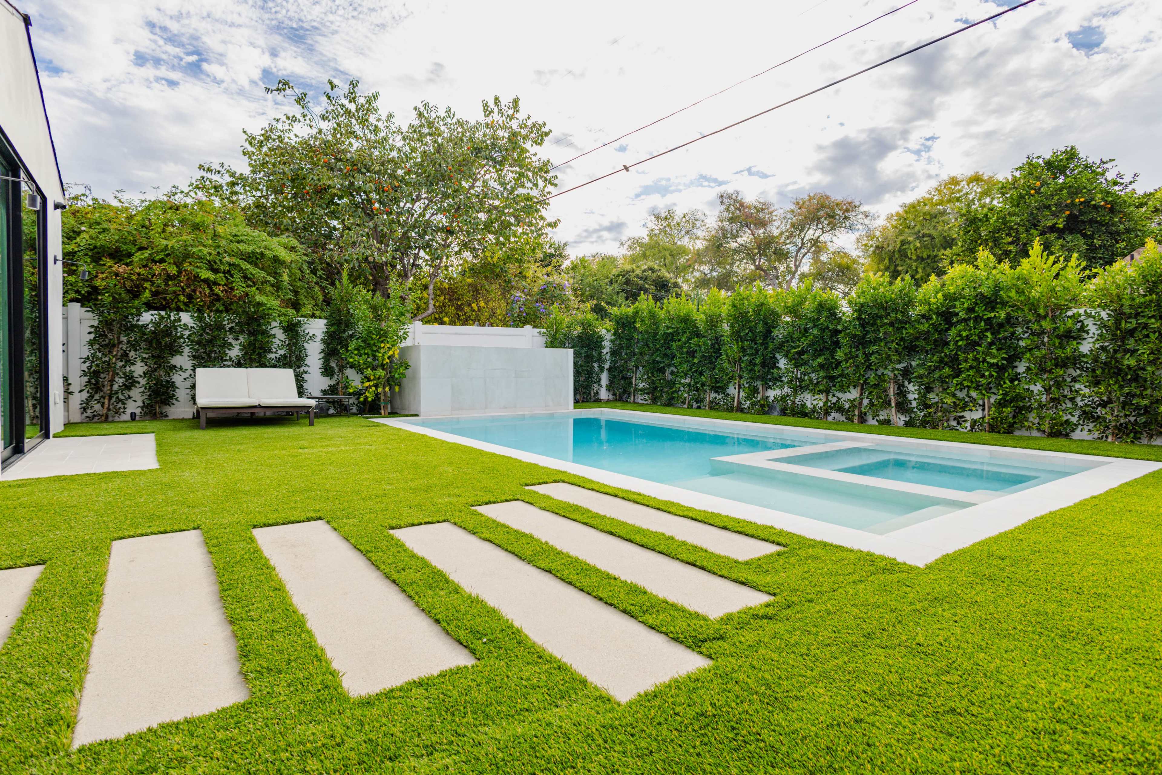 The image shows a modern backyard featuring a swimming pool surrounded by manicured grass and concrete stepping stones.
