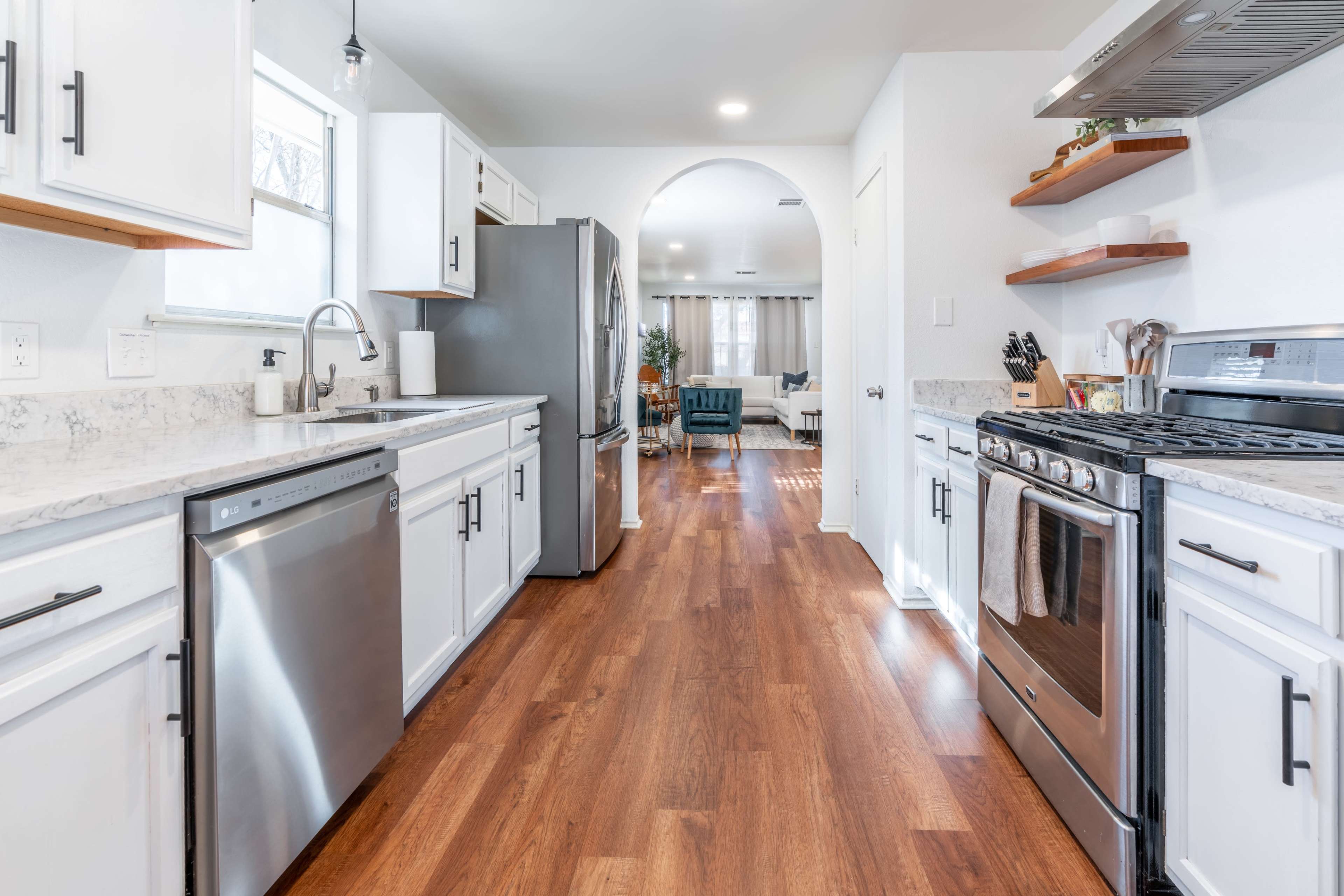A modern kitchen features stainless steel appliances, white cabinetry, and hardwood flooring, with an open archway leading to a living area.