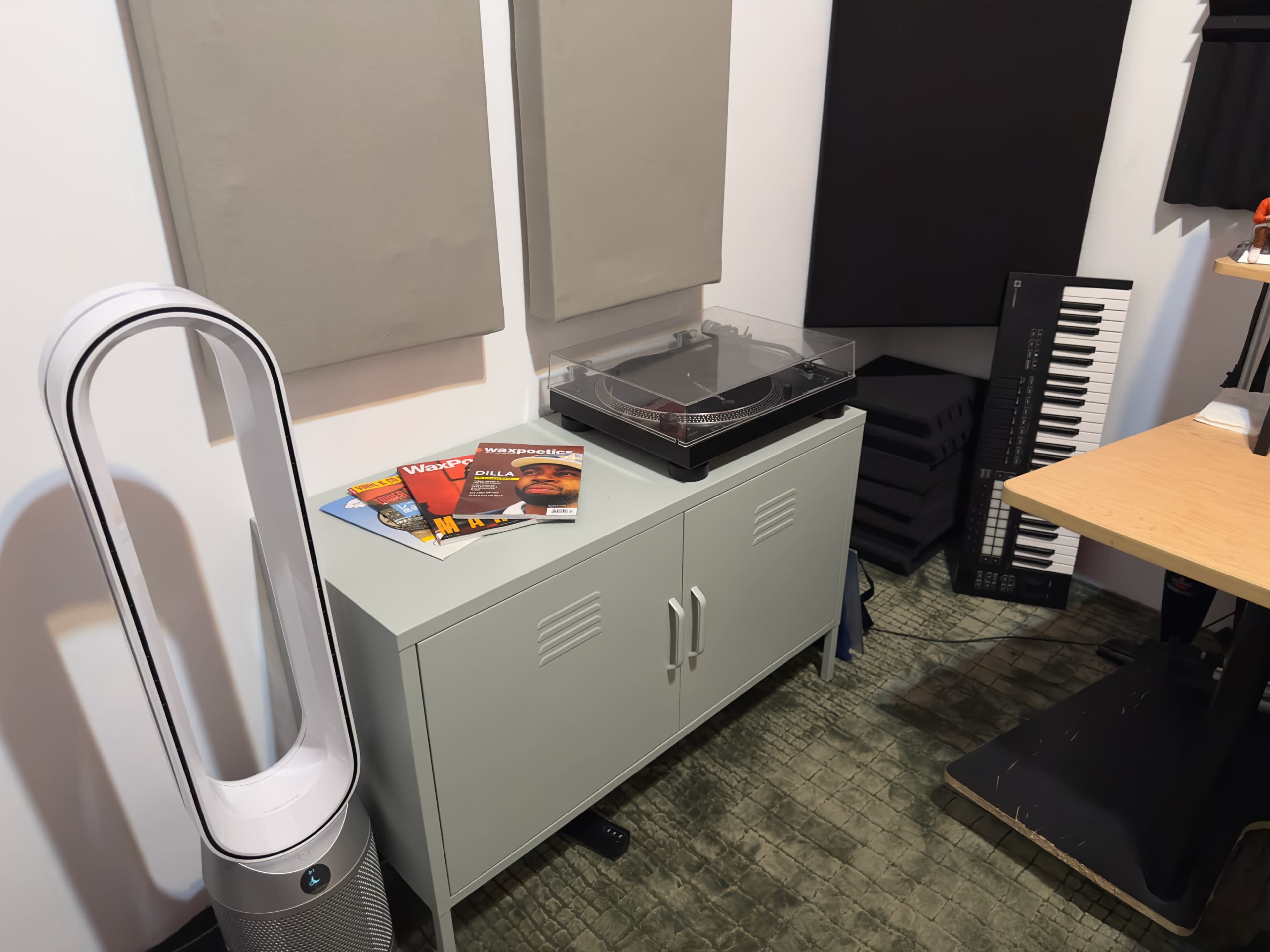 The image shows a compact room featuring a gray cabinet with magazines on top, a turntable, a keyboard, and fabric soundproofing panels on the walls.