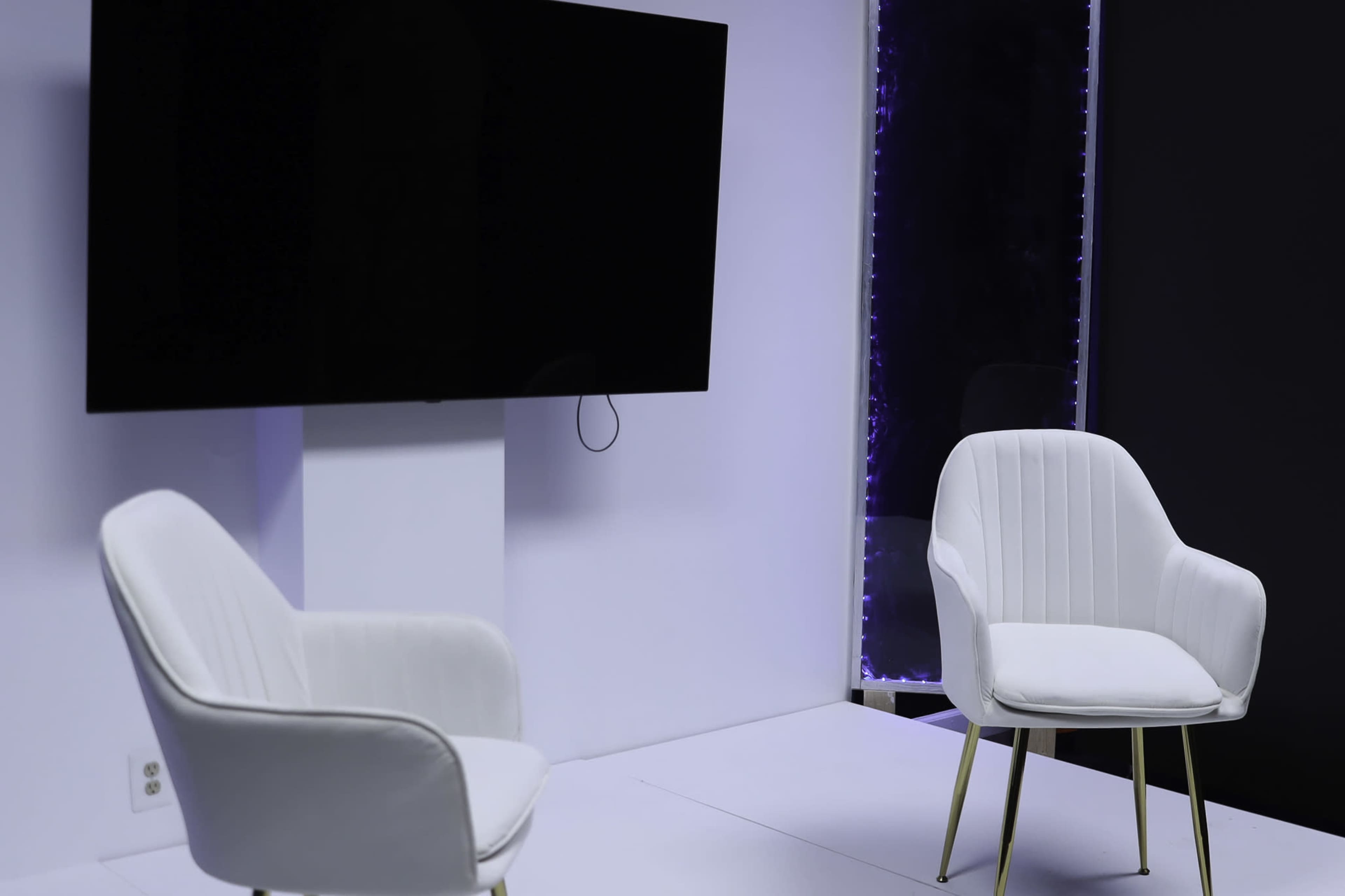 The image shows a minimalist interview setting featuring two white chairs facing a large black television screen on a light-colored backdrop.