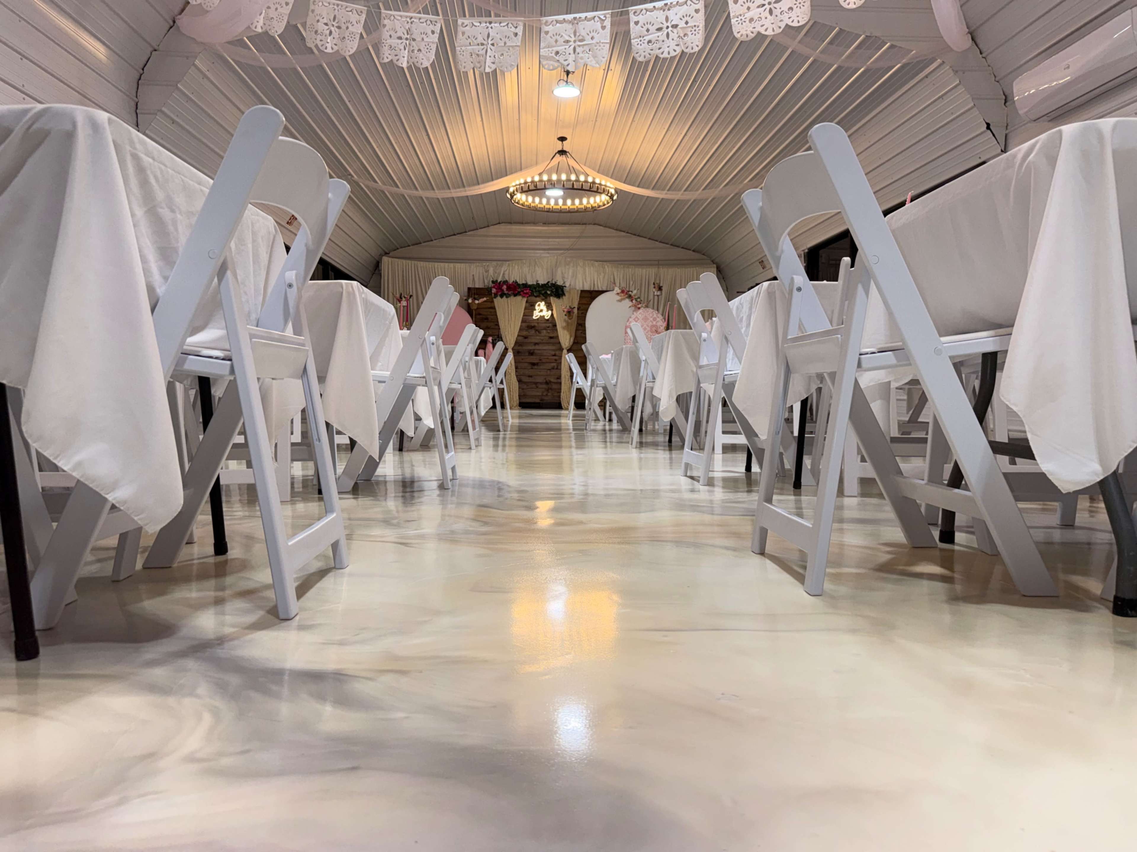 The image shows a dining area with white tables covered in cloth, lined with folding chairs beneath a decorative ceiling and a chandelier.