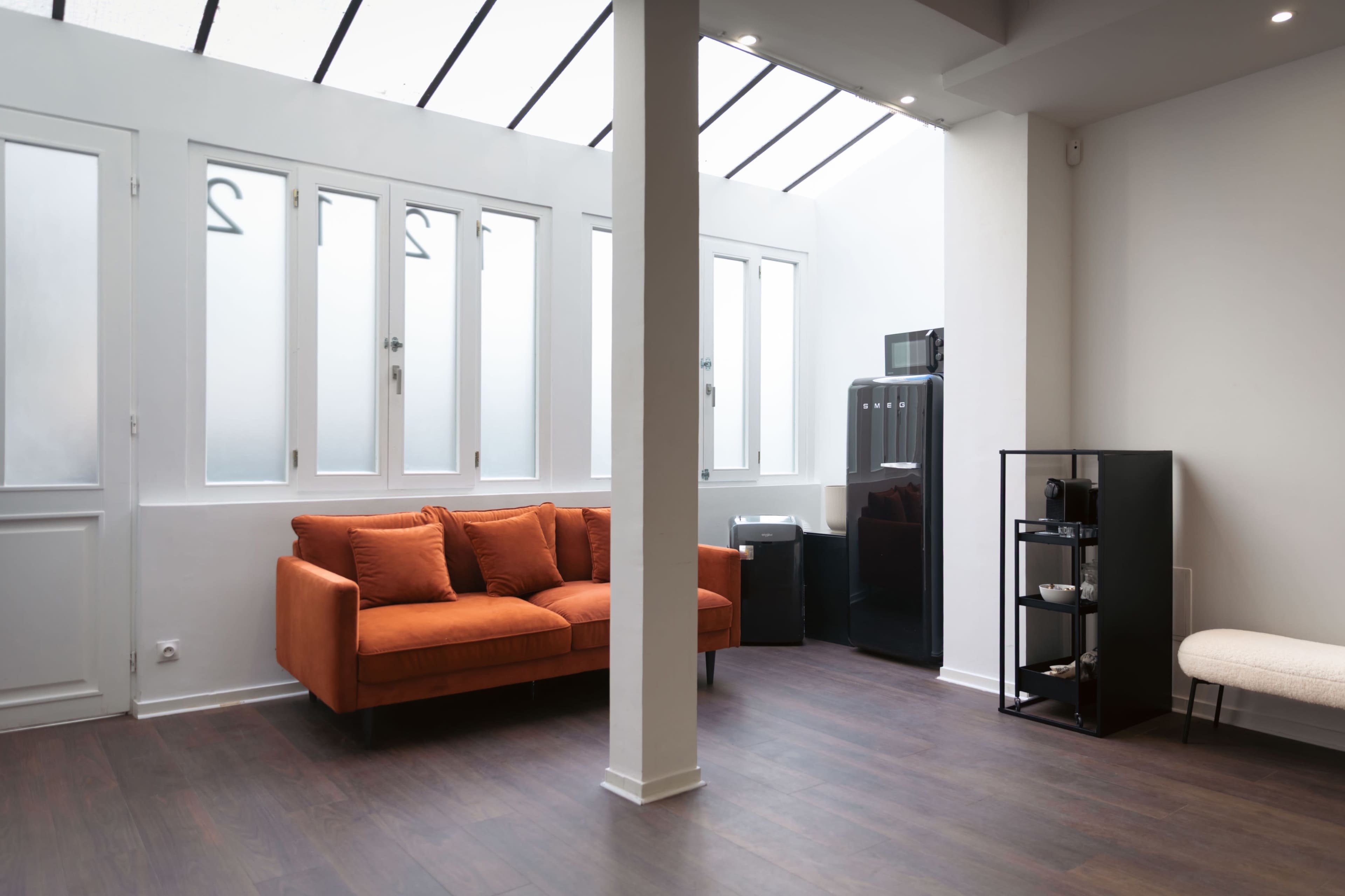 The image shows a modern, minimalist room featuring an orange sofa, a black refrigerator, and a shelving unit against white walls with large windows.