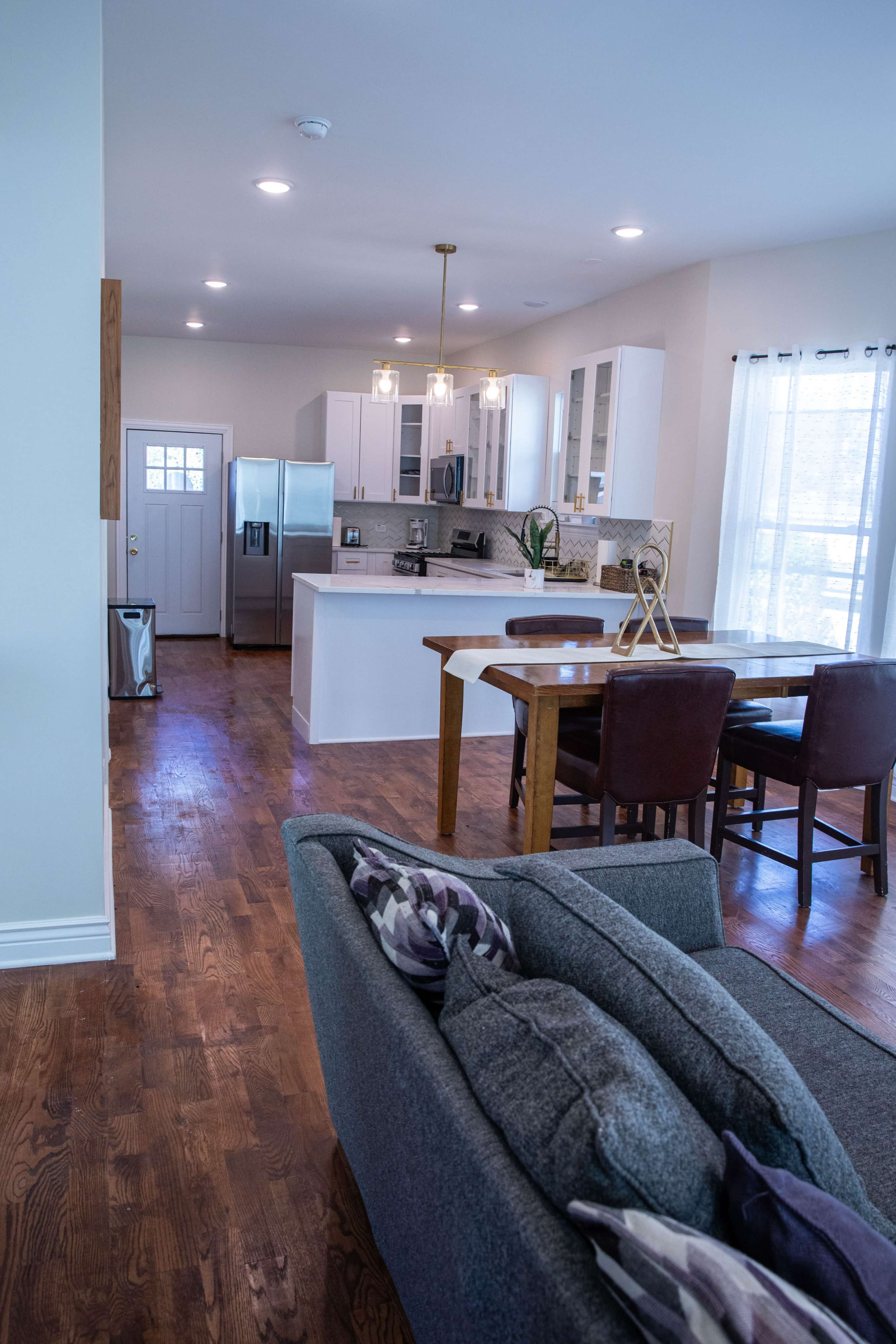 A modern kitchen and dining area features a wooden table with chairs, stainless steel appliances, and pendant lighting, visible from a cozy living room with a sofa.