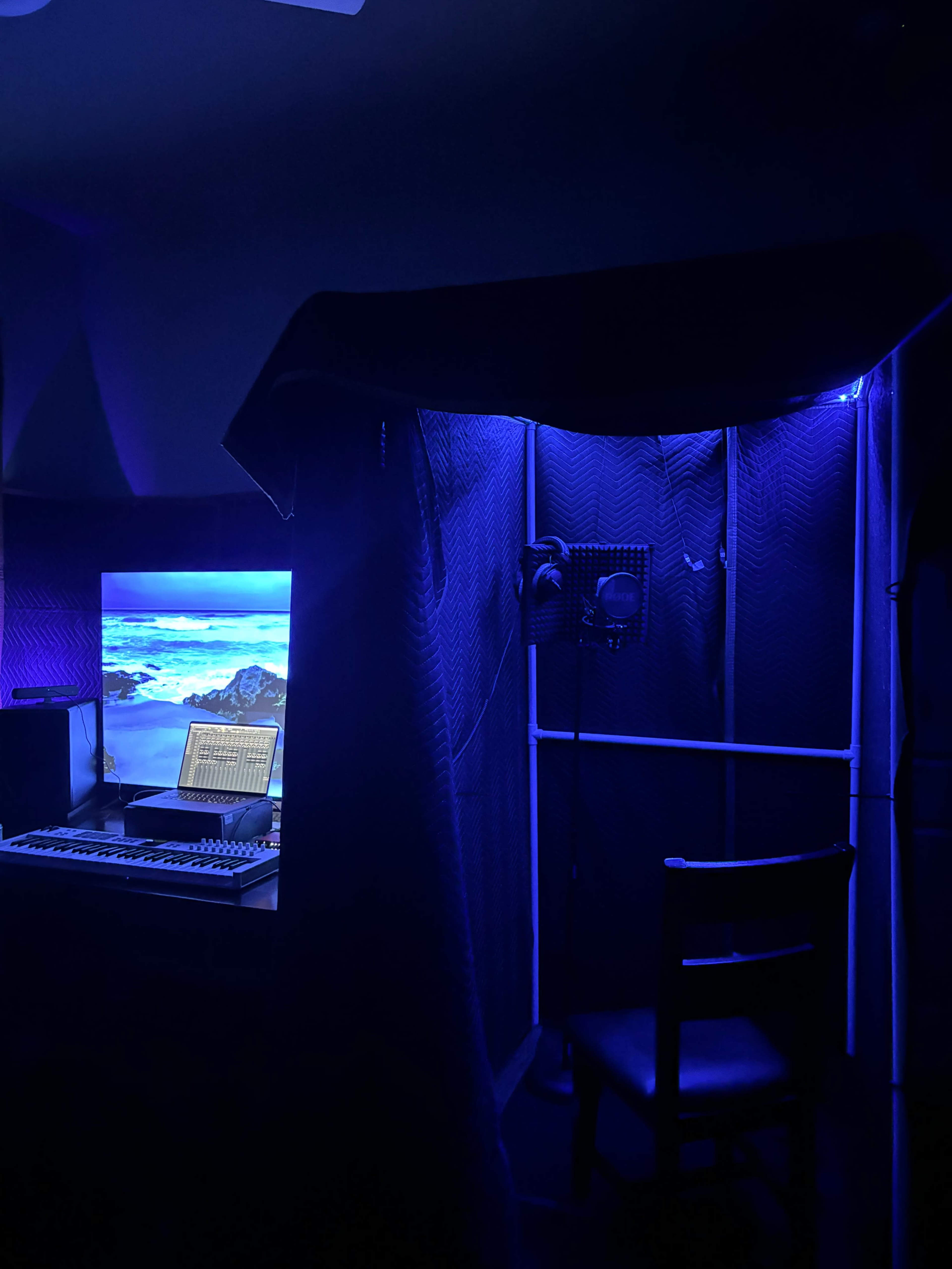 A dimly lit recording studio features a computer setup with a monitor displaying a scenic image, surrounded by soundproofing panels and a microphone on a stand.