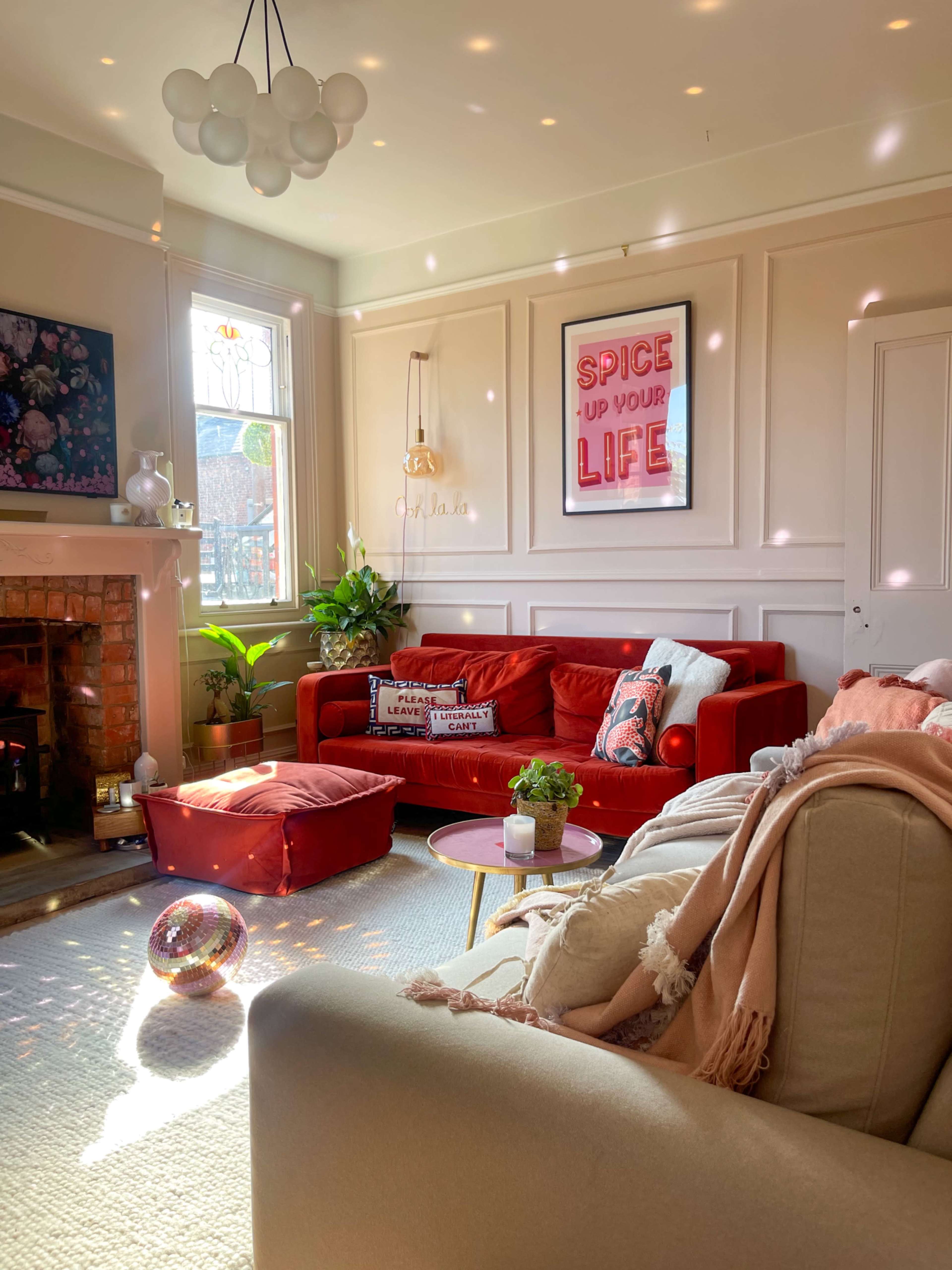 The bright living room features a red sofa, a light-colored armchair, and a fireplace, with decorative plants and a colorful framed poster on the wall.