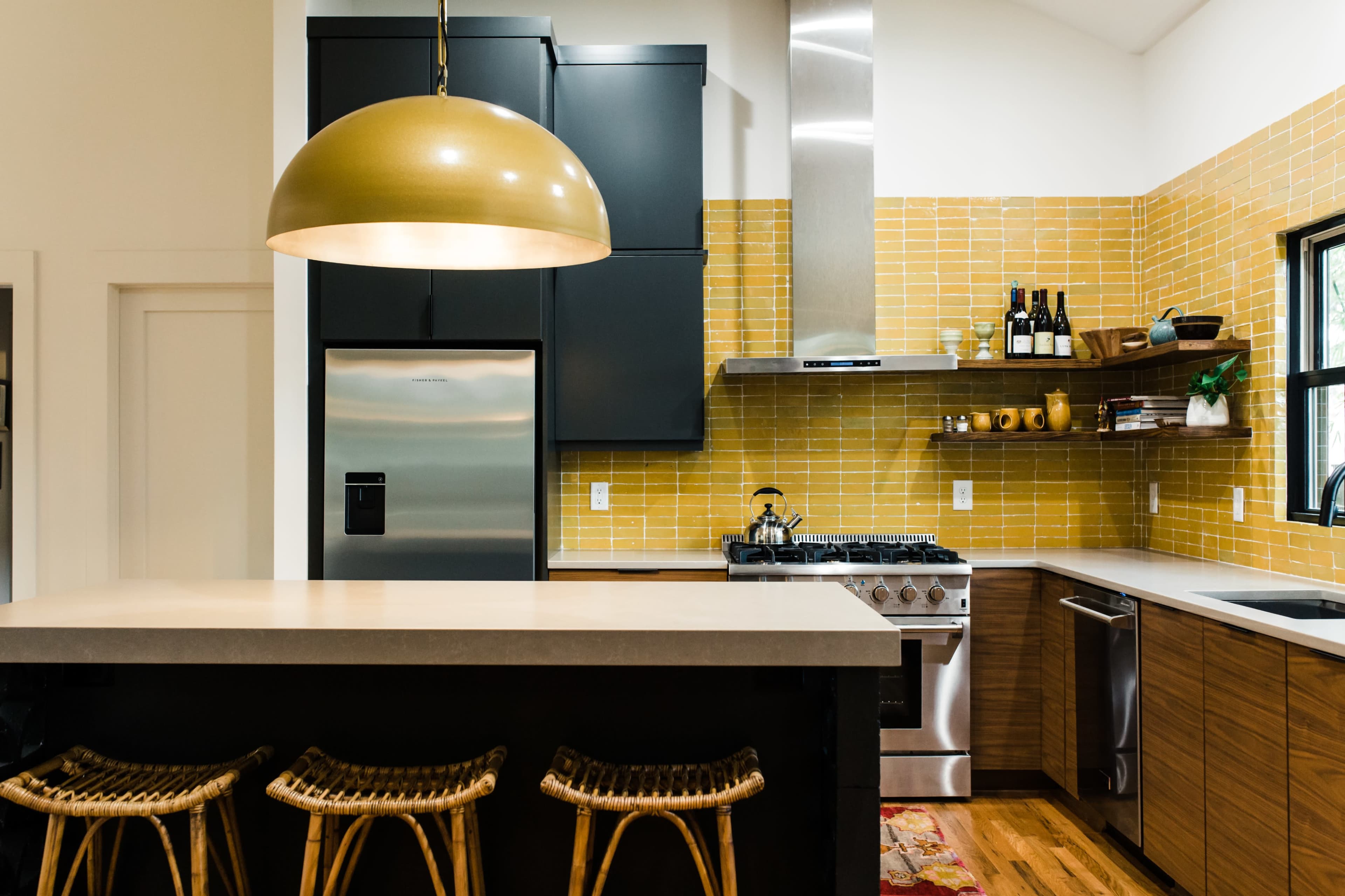 A modern kitchen features a large pendant light, stainless steel appliances, and yellow tiled walls above wooden cabinetry.