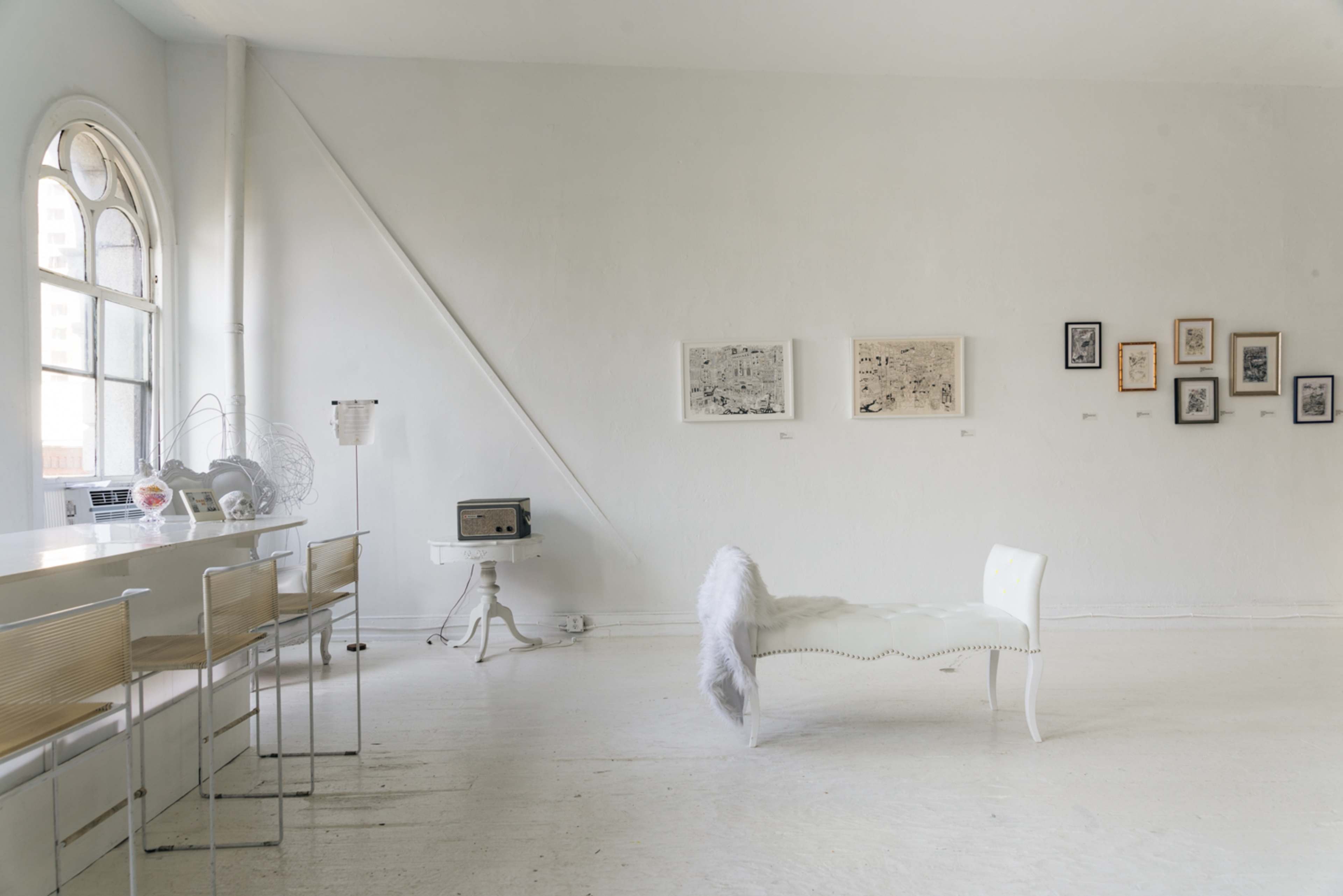 A bright, minimalist art gallery features white walls, a bench with a fur throw, and several framed artworks hanging on the walls.