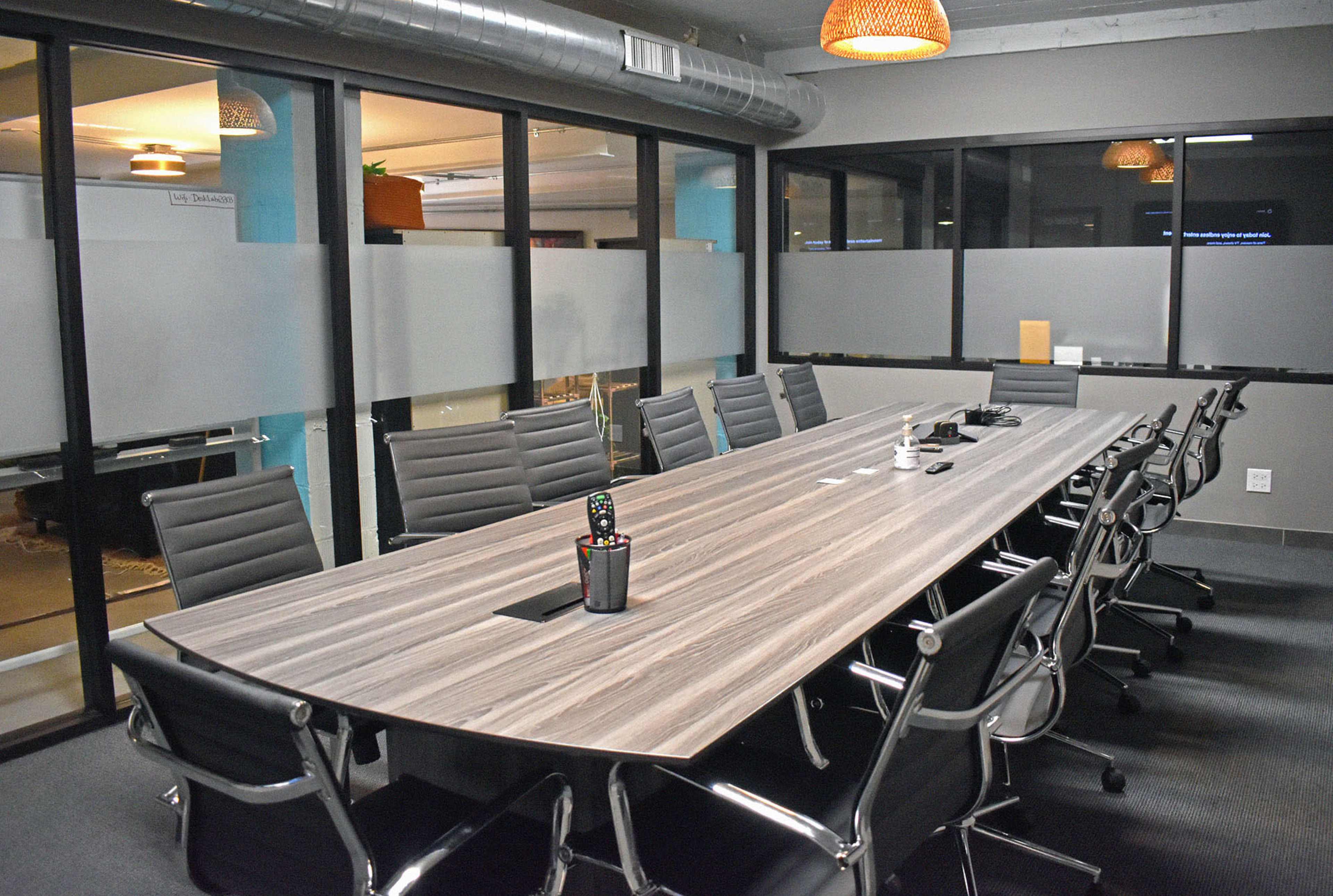 A large conference room features a long wooden table surrounded by ergonomic chairs, equipped with a remote control and other office supplies on the table.