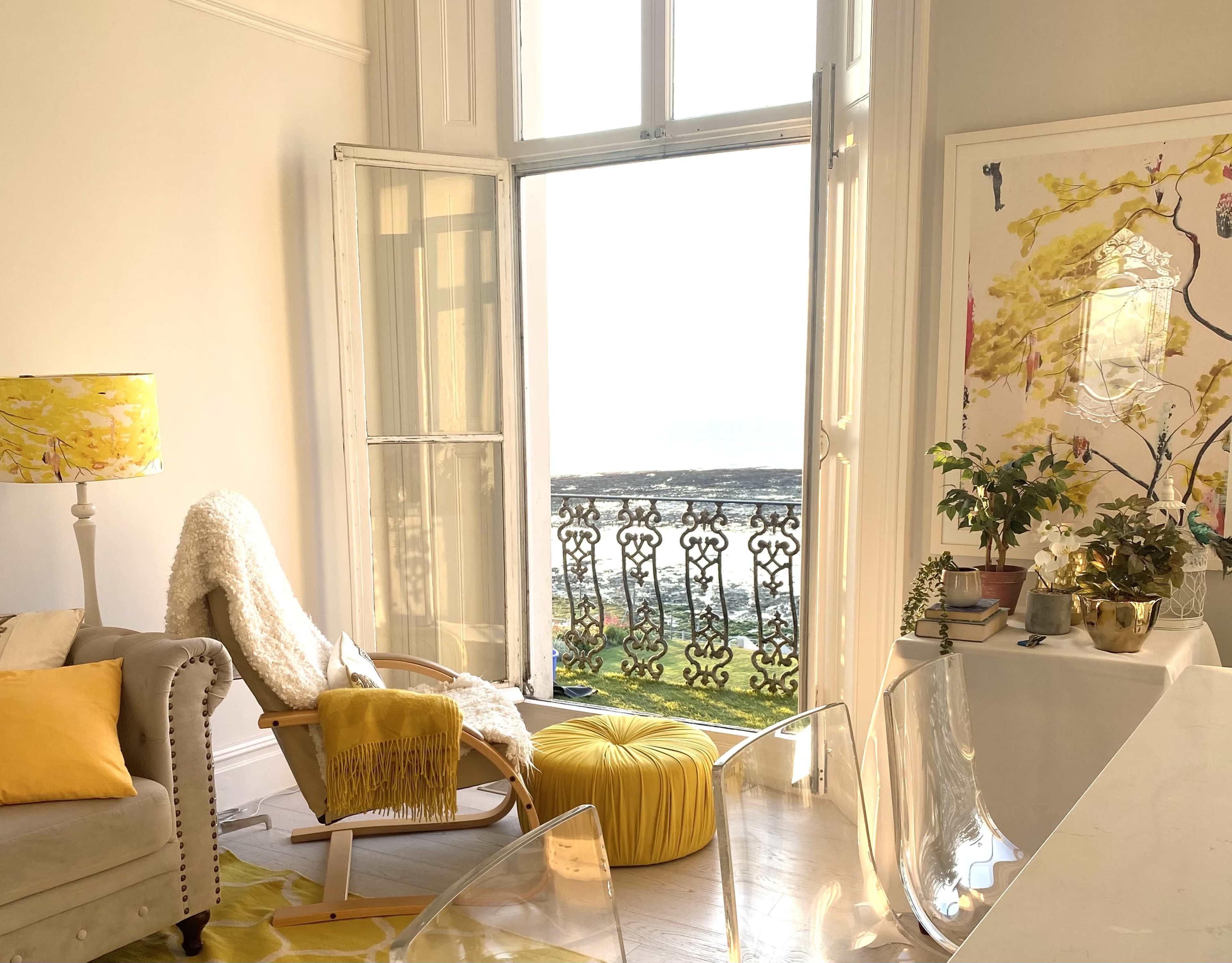 A bright living room features a large window with open doors overlooking a scenic view, complemented by a comfortable sofa, a yellow pouf, and decorative plant arrangements.