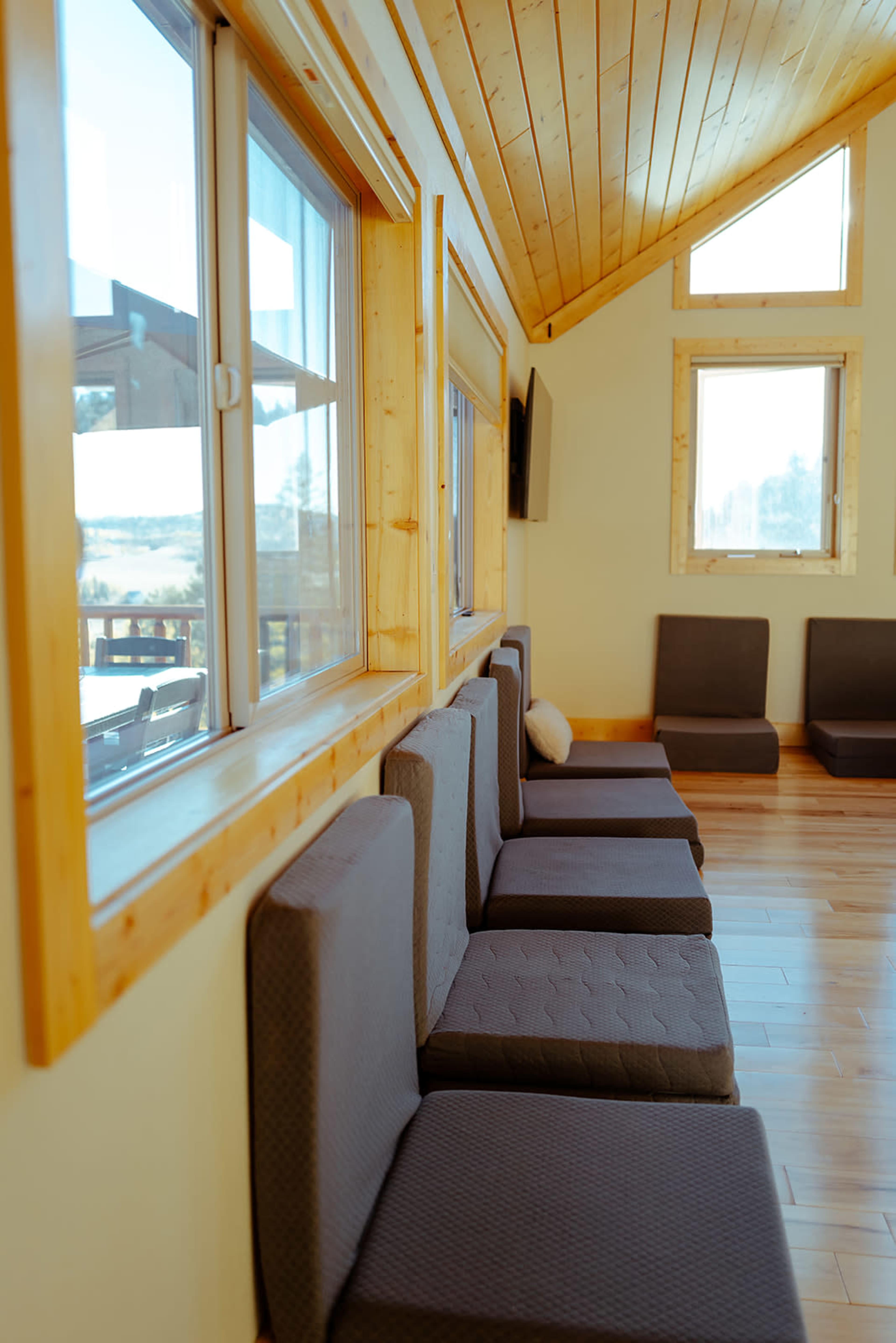 The image shows a row of simple, upholstered seating against a wooden wall with large windows and a high wooden ceiling.