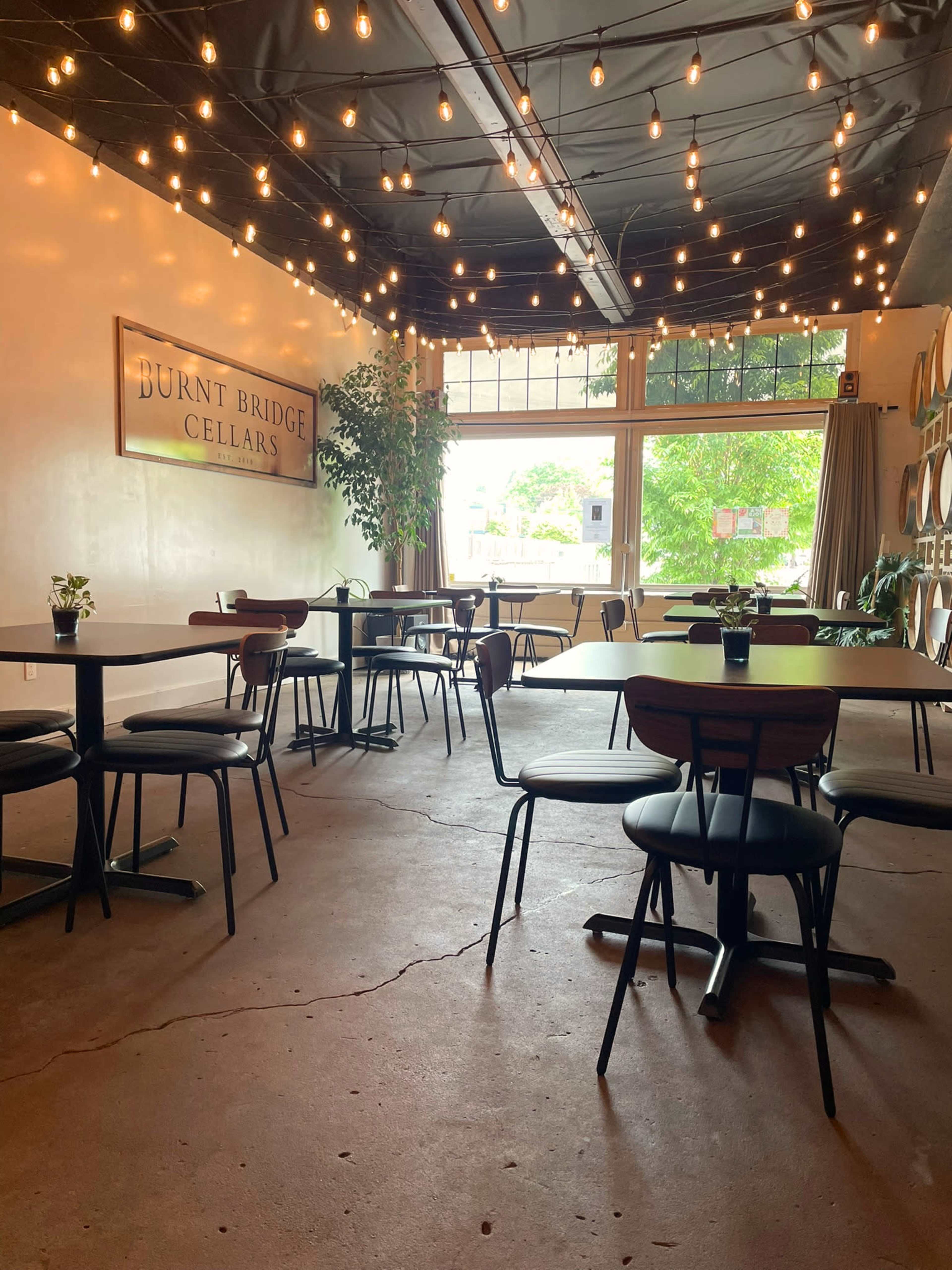 The interior of a wine cellar features several wooden tables and chairs under decorative string lights, with large windows allowing natural light to illuminate the space.