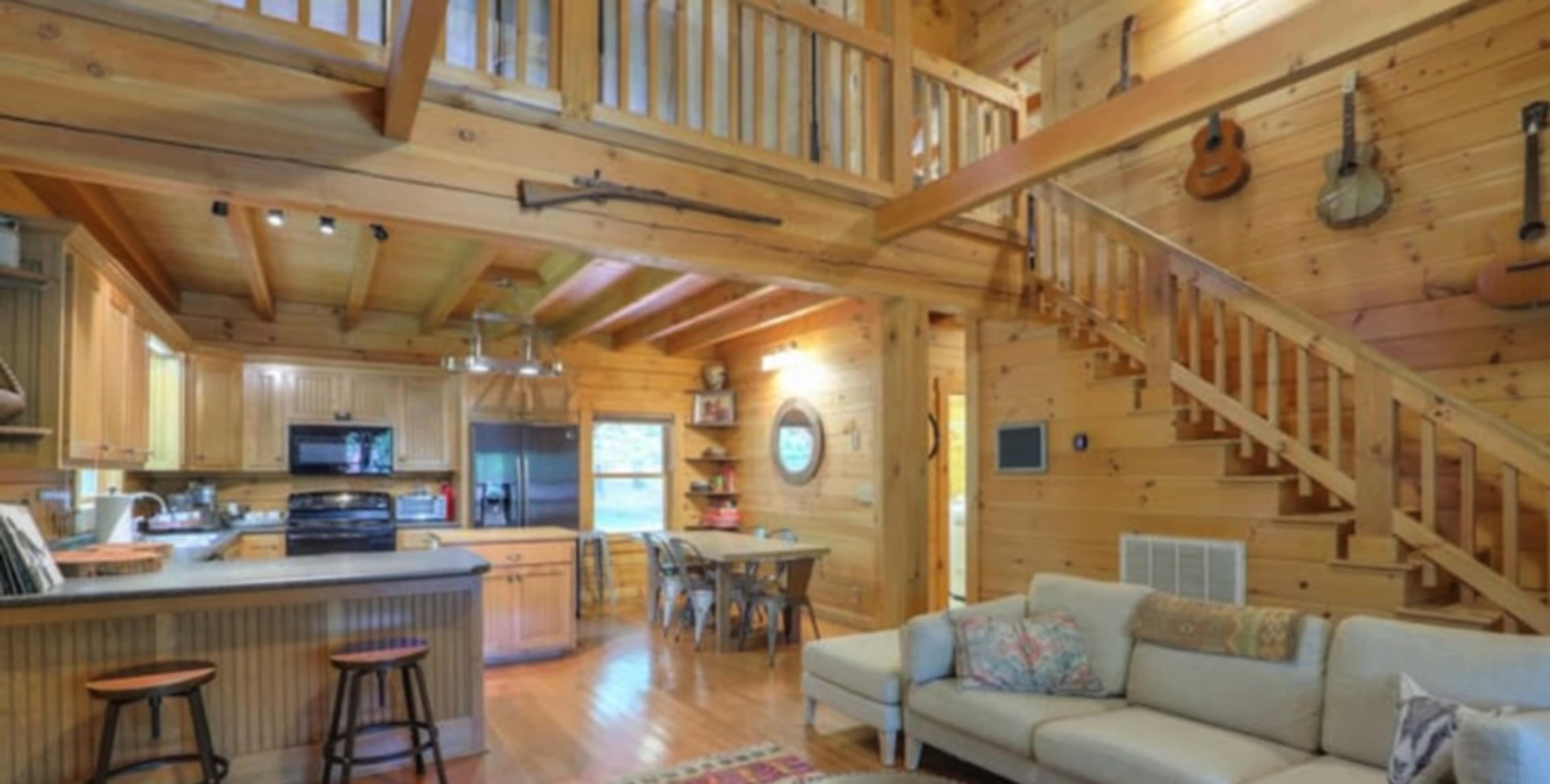A wooden cabin interior featuring an open layout with a kitchen, dining area, and a living space, along with a staircase leading to a loft.