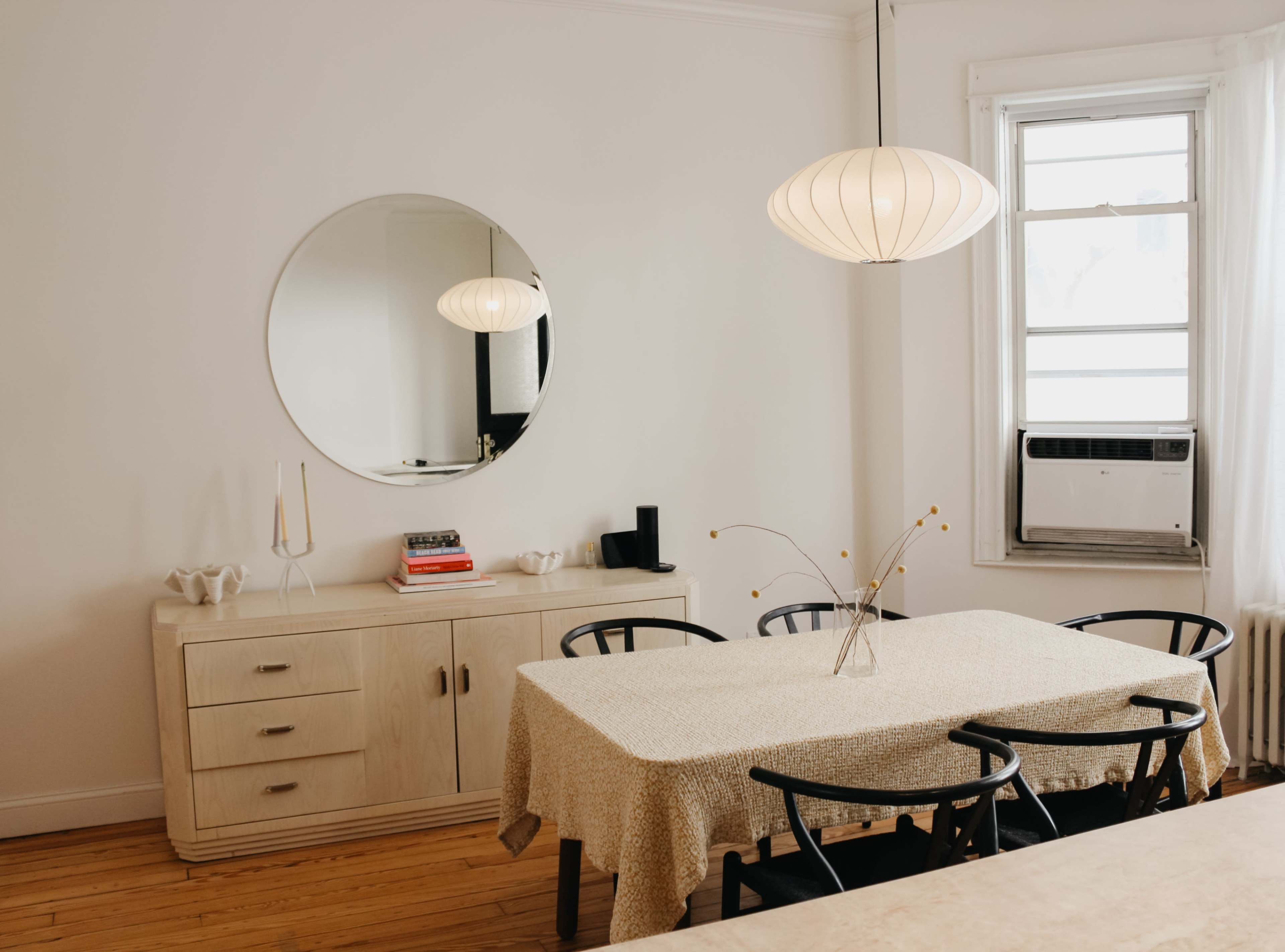 A dining area features a round table with a beige tablecloth, surrounded by black chairs, under a pendant light, with a large mirror and a window in the background.
