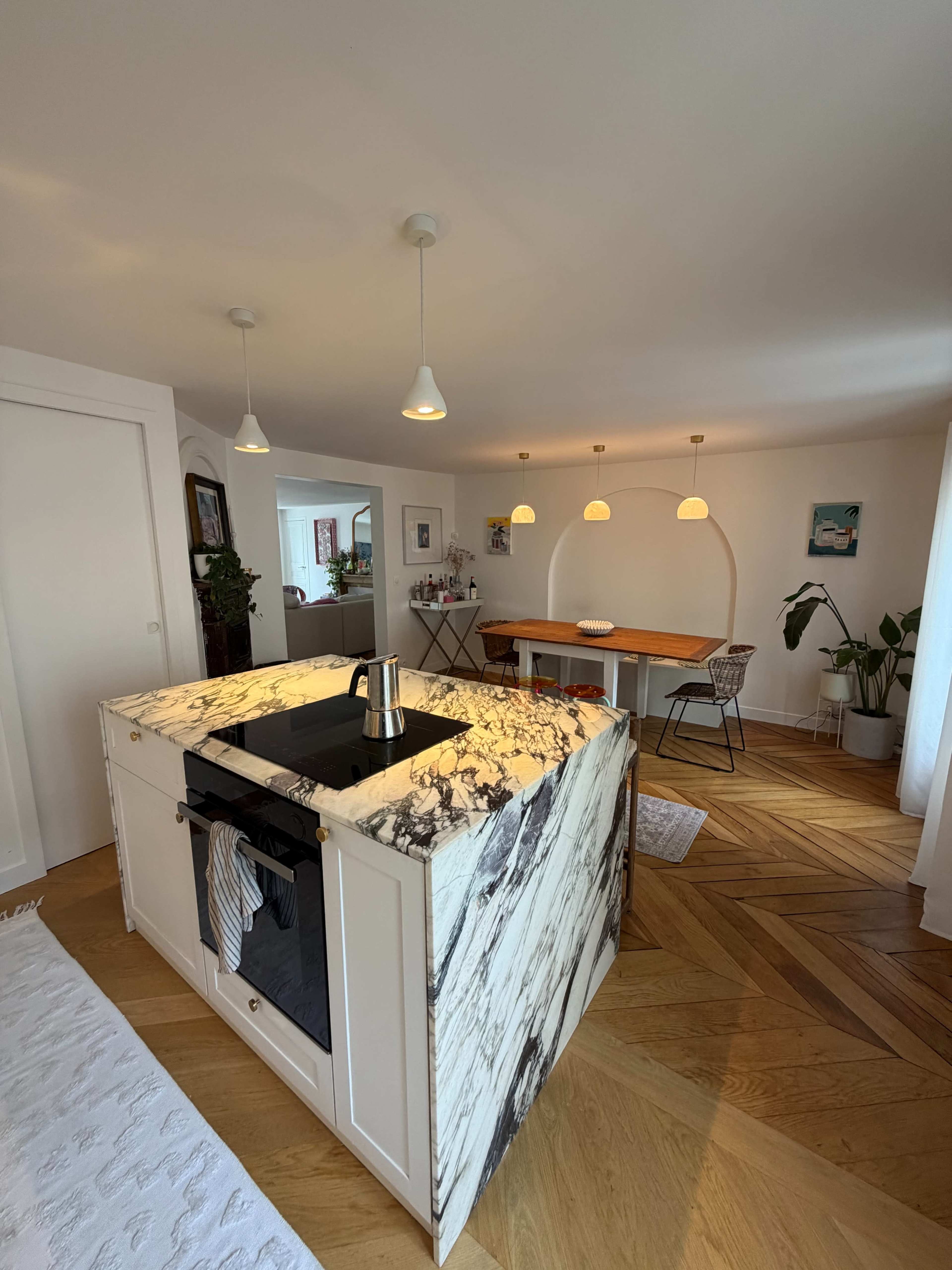 The image shows a modern kitchen with a marble island and a dining area featuring a wooden table illuminated by three pendant lights.