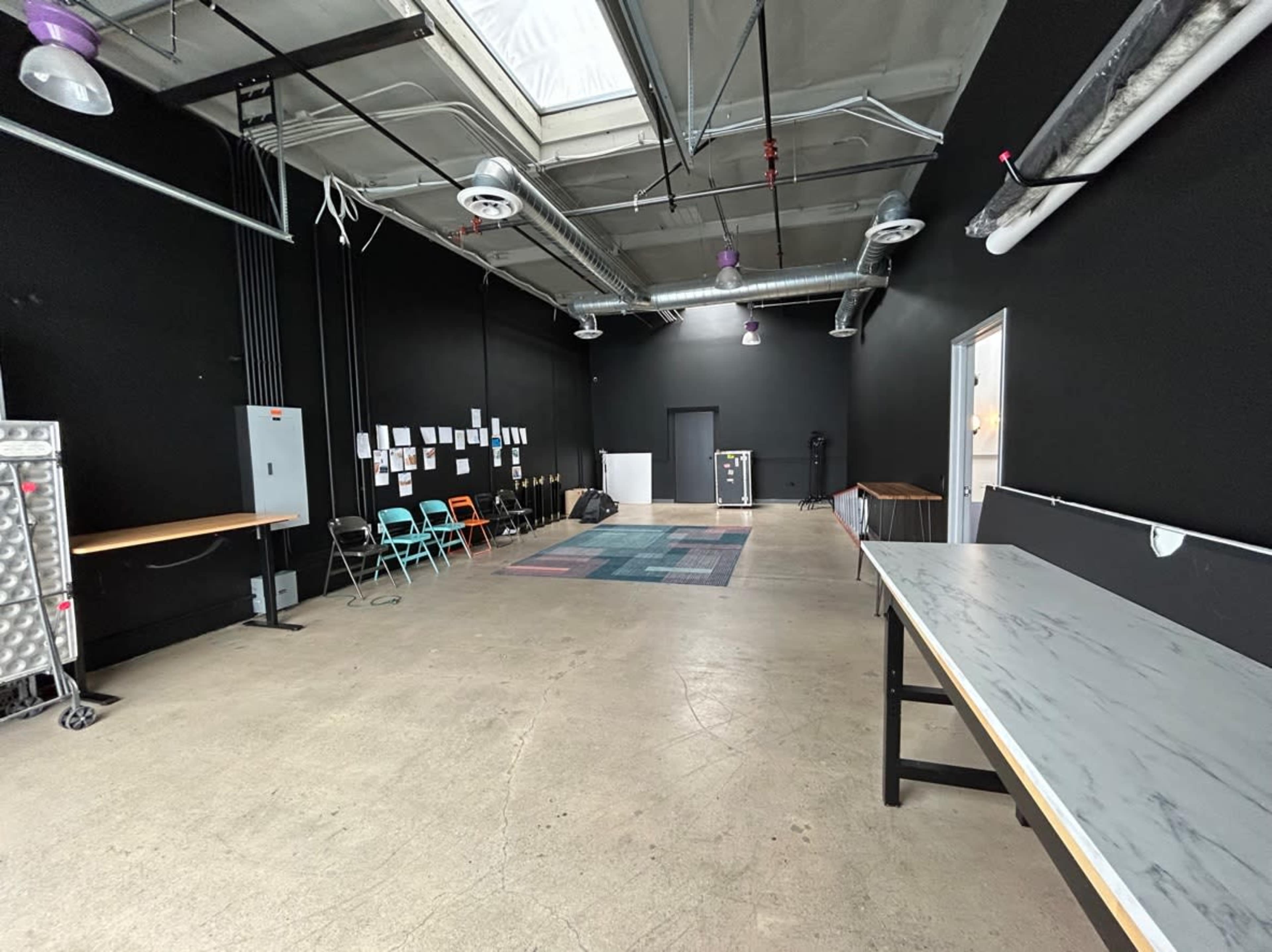 An empty room with black walls, light concrete flooring, and several tables along the sides, with a carpet and a few chairs in the center.