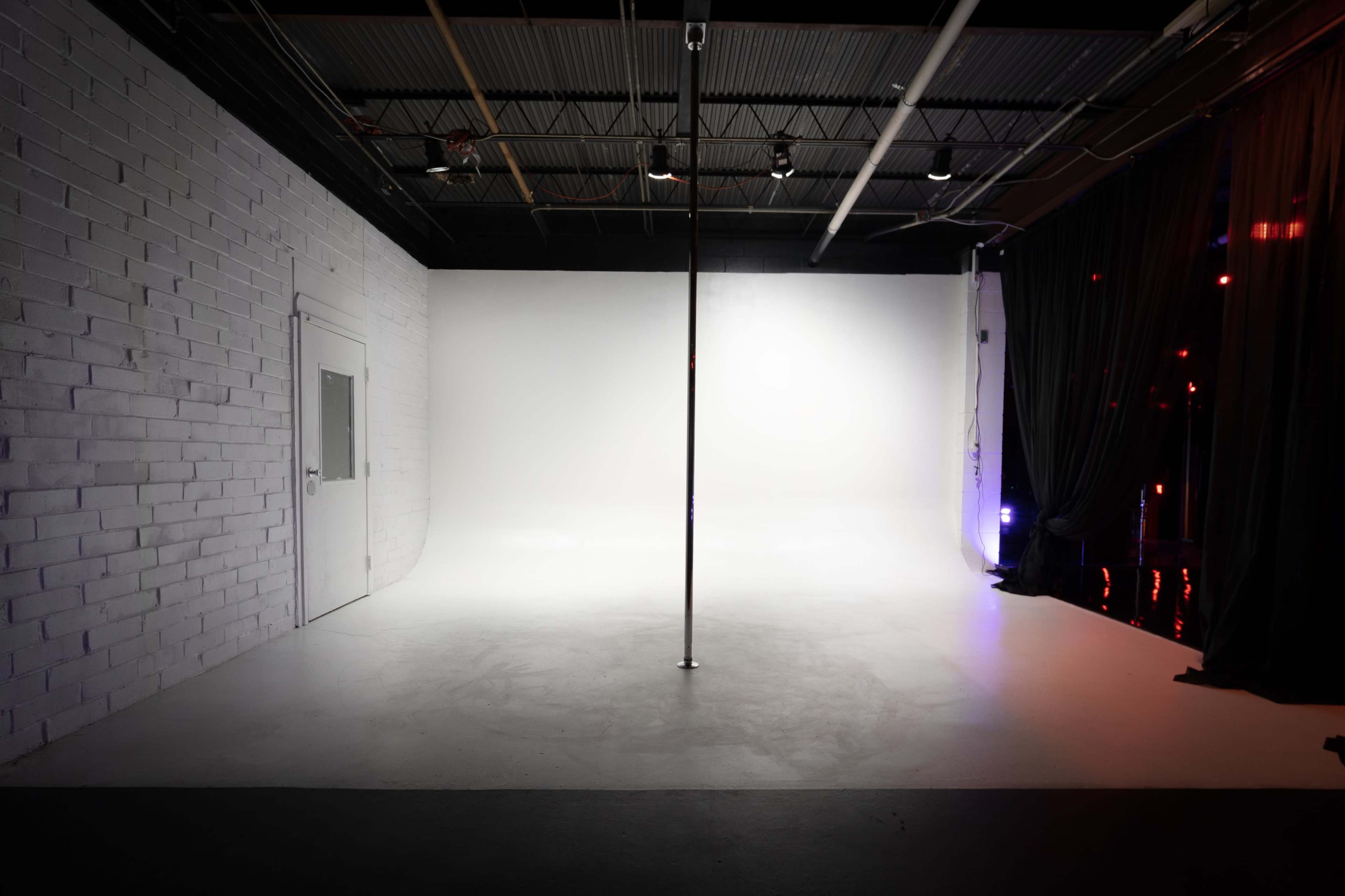The image shows an empty studio space with a white backdrop, a central pole, and dim lighting.