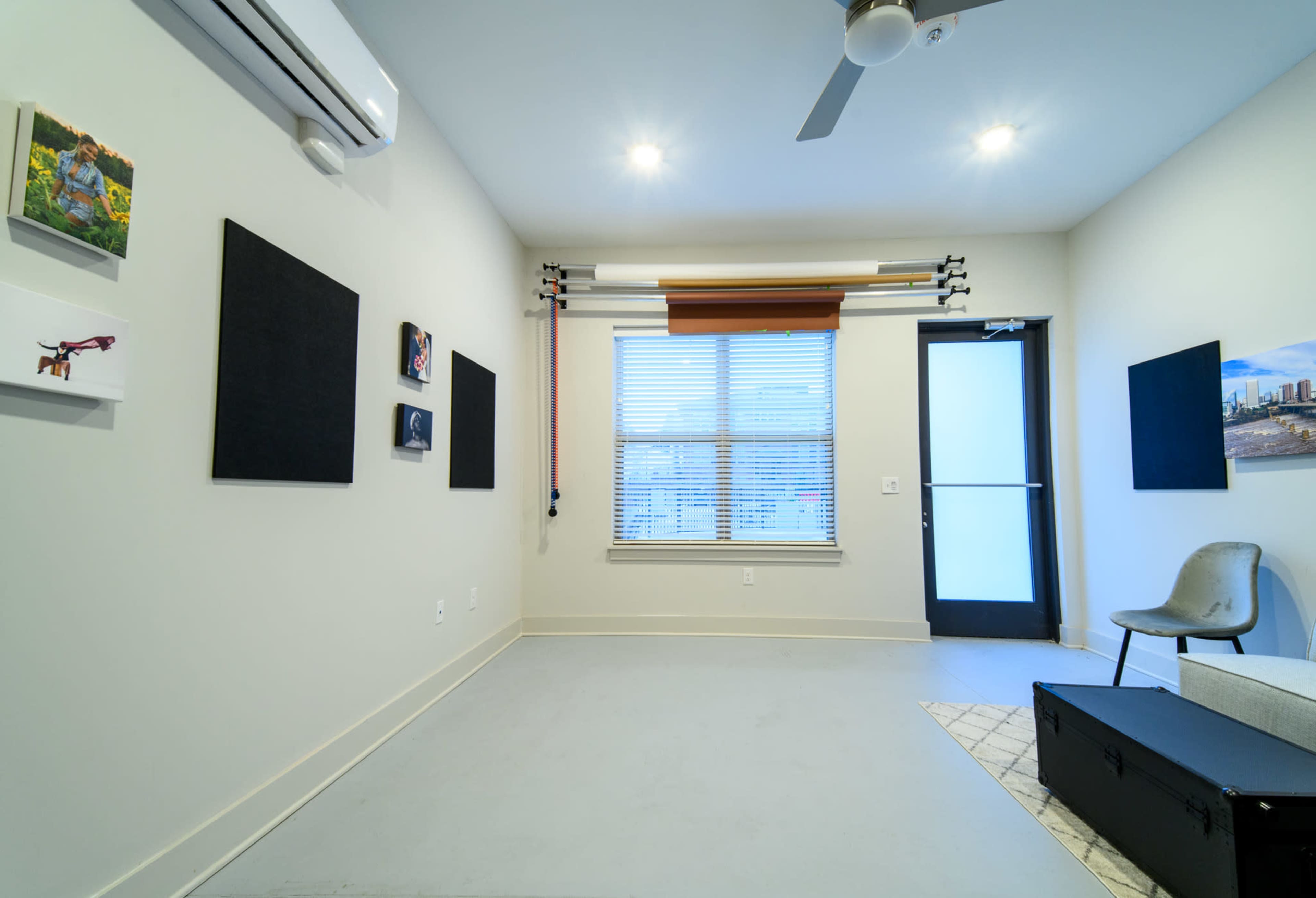 The image shows a sparsely furnished room with two black wall panels, a window with blinds, and a chair near the door.