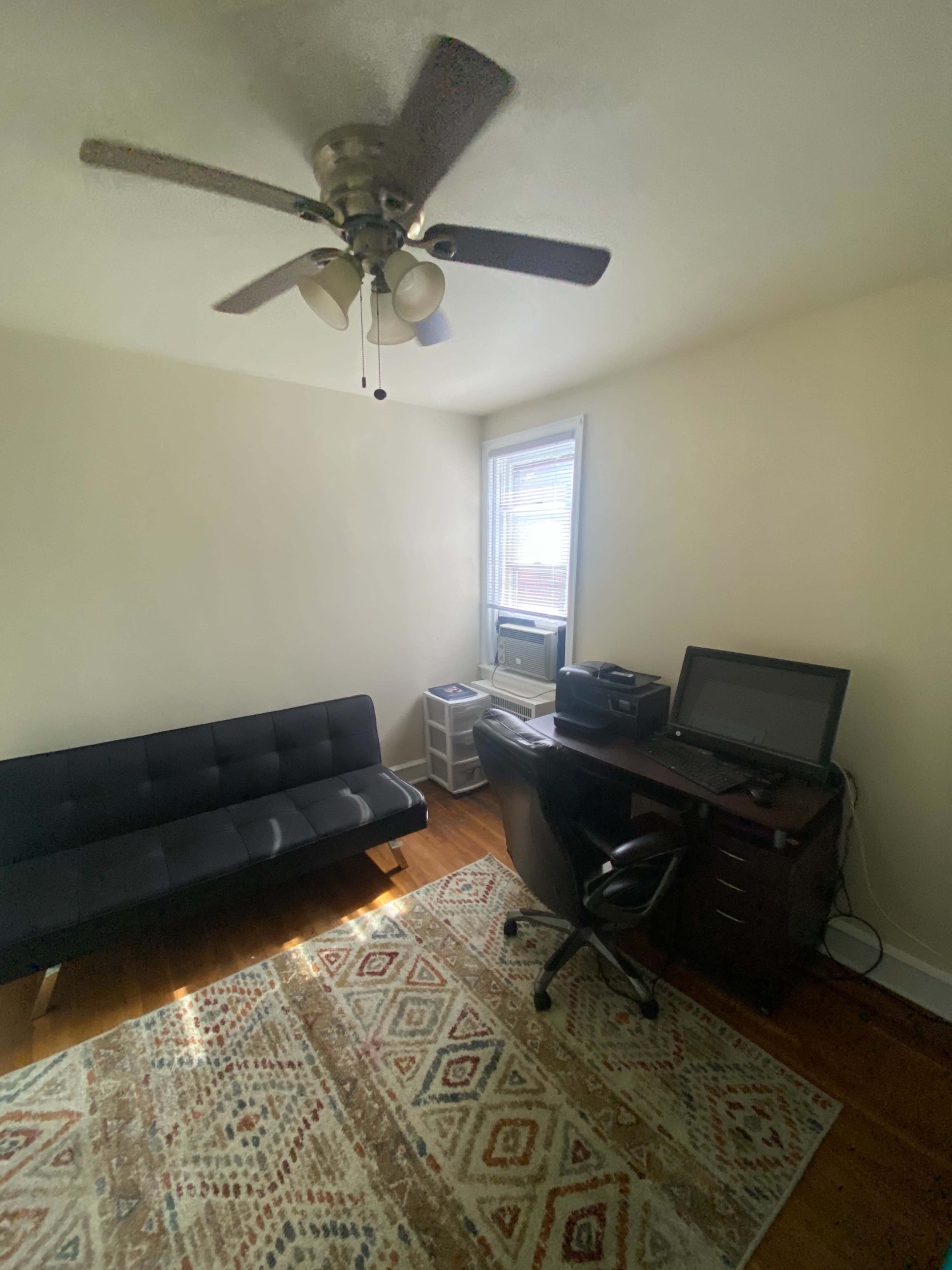 The image shows a small room with a futon, a desk with a computer, and a window allowing natural light to enter.