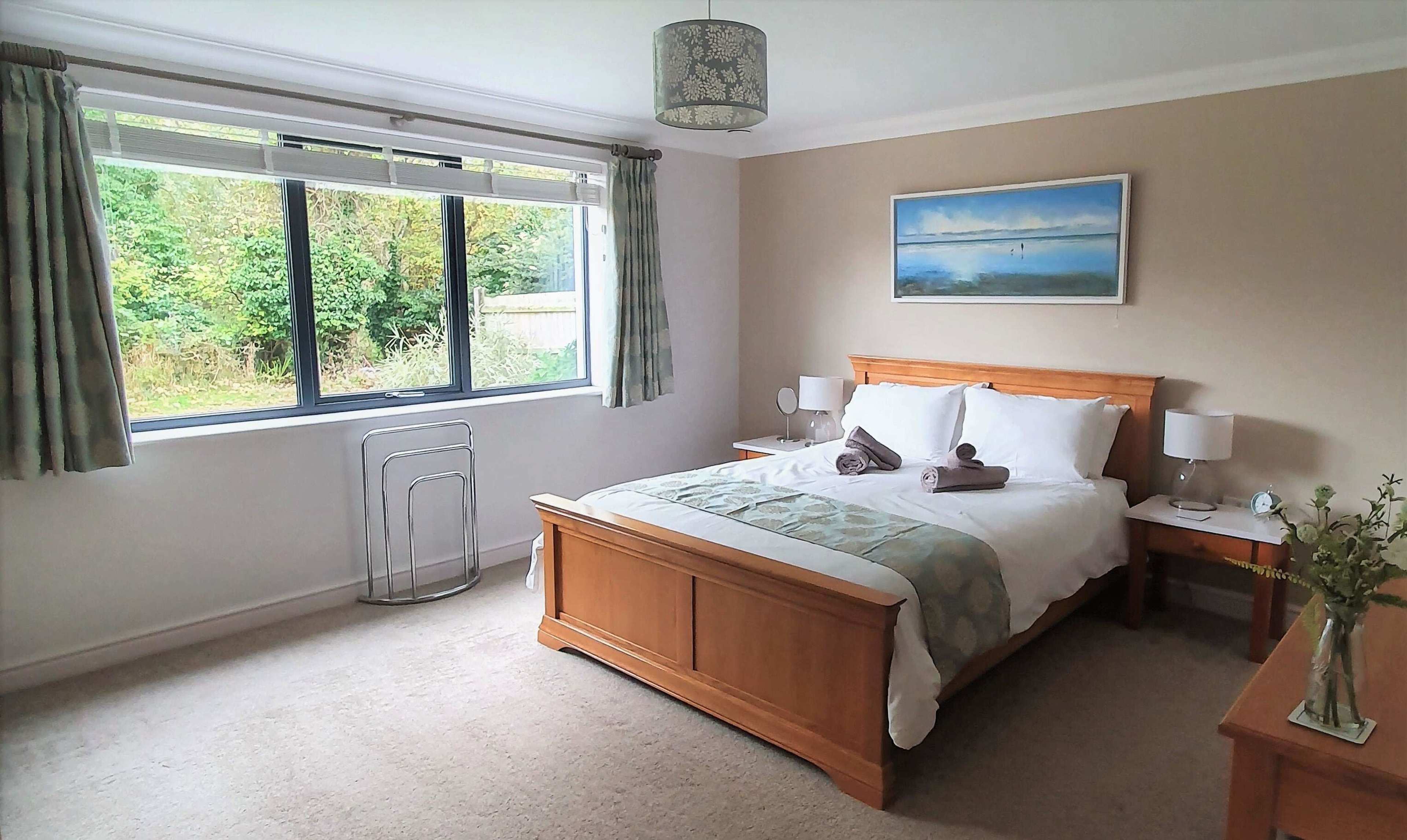 A tidy bedroom features a wooden bed with white bedding, nightstands with lamps, and a large window overlooking greenery.